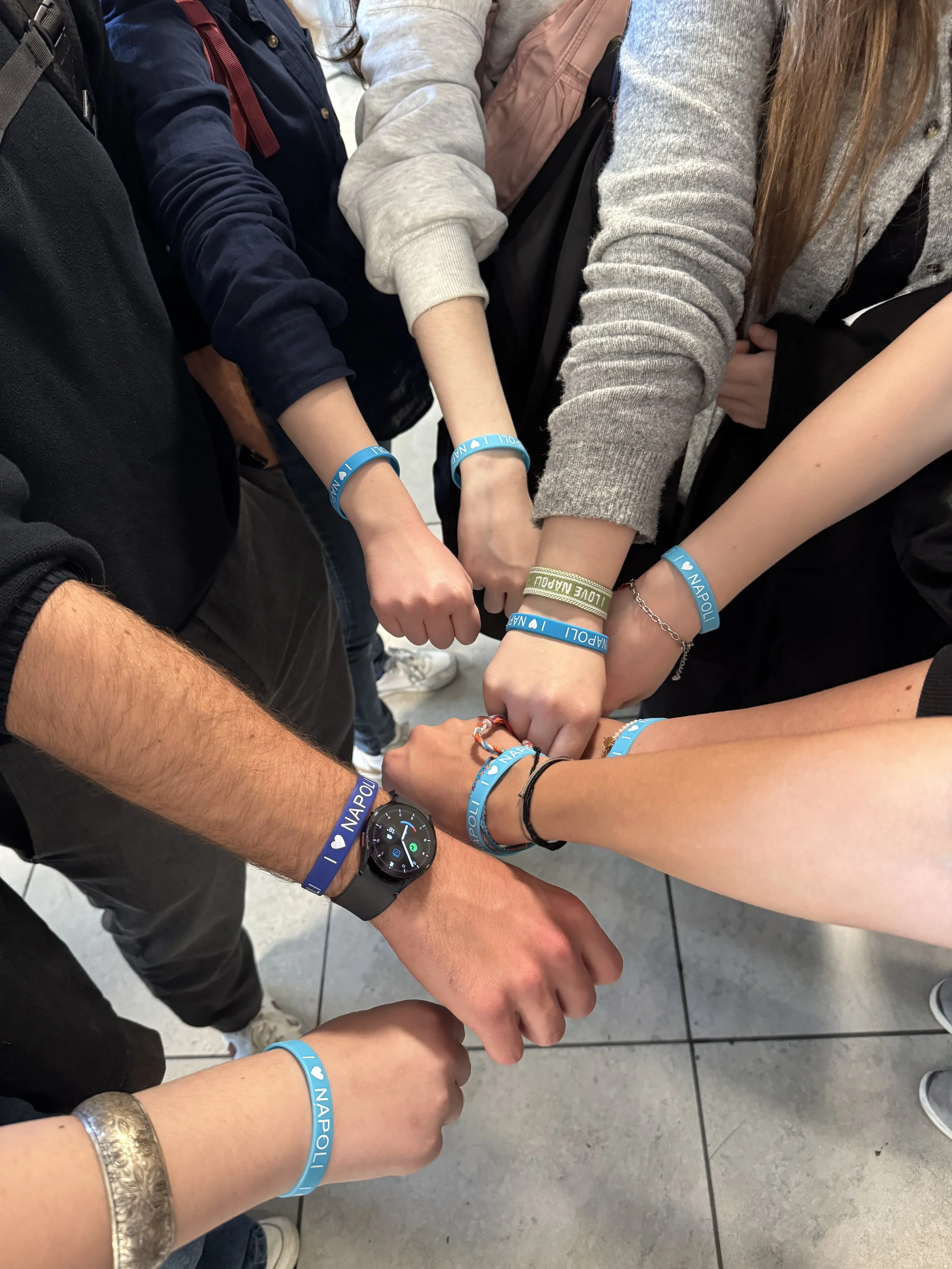 Mehrere Personen zeigen ihre Handgelenke mit blauen Armbändern, die den Text 'I ♥ NAPOLI' tragen, und einer Person trägt eine Smartwatch.