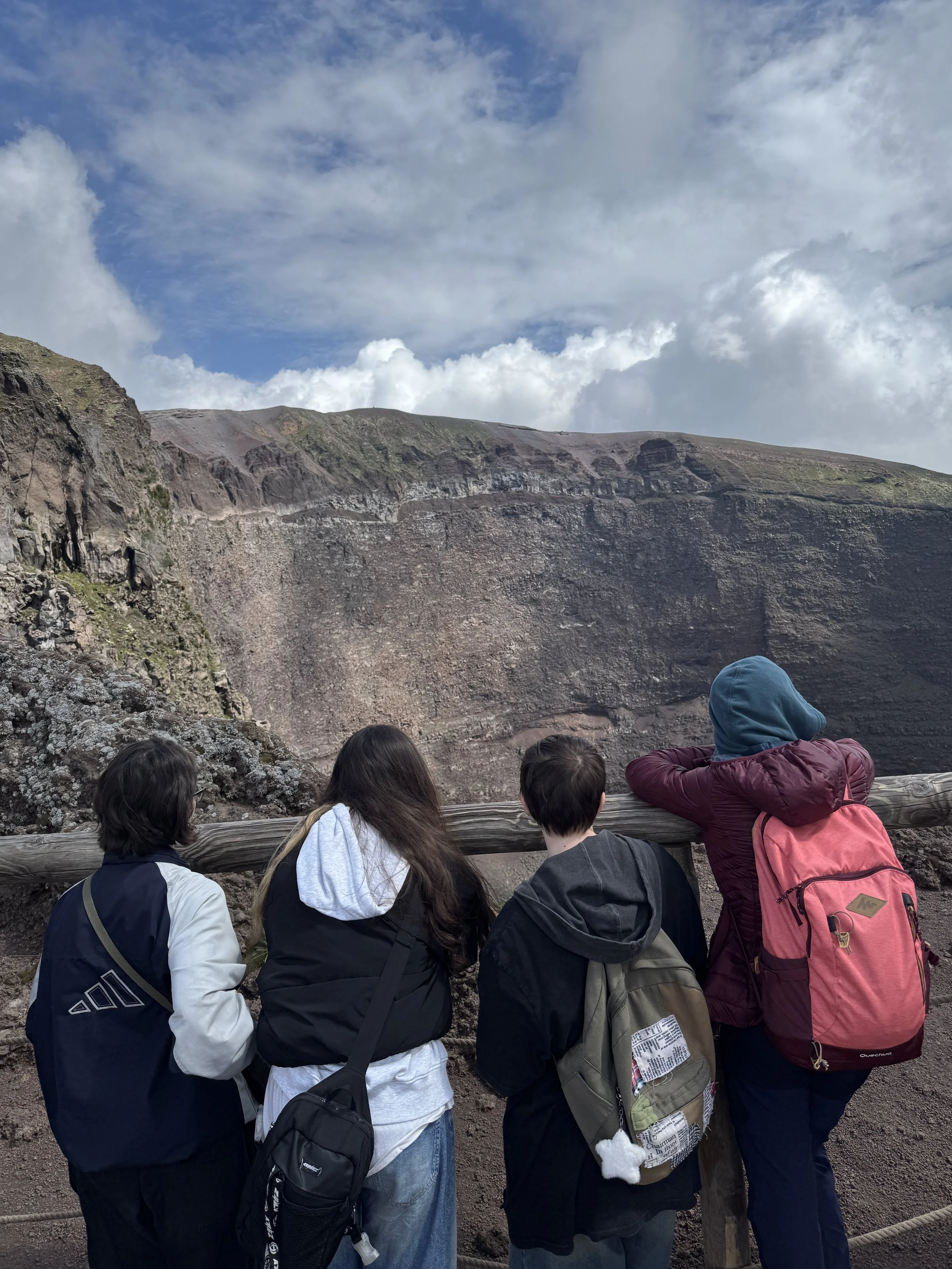 Vier Jugendliche stehen am Zaun und schauen auf einen Krater eines Vulkans, mit Himmel und Wolken im Hintergrund.
