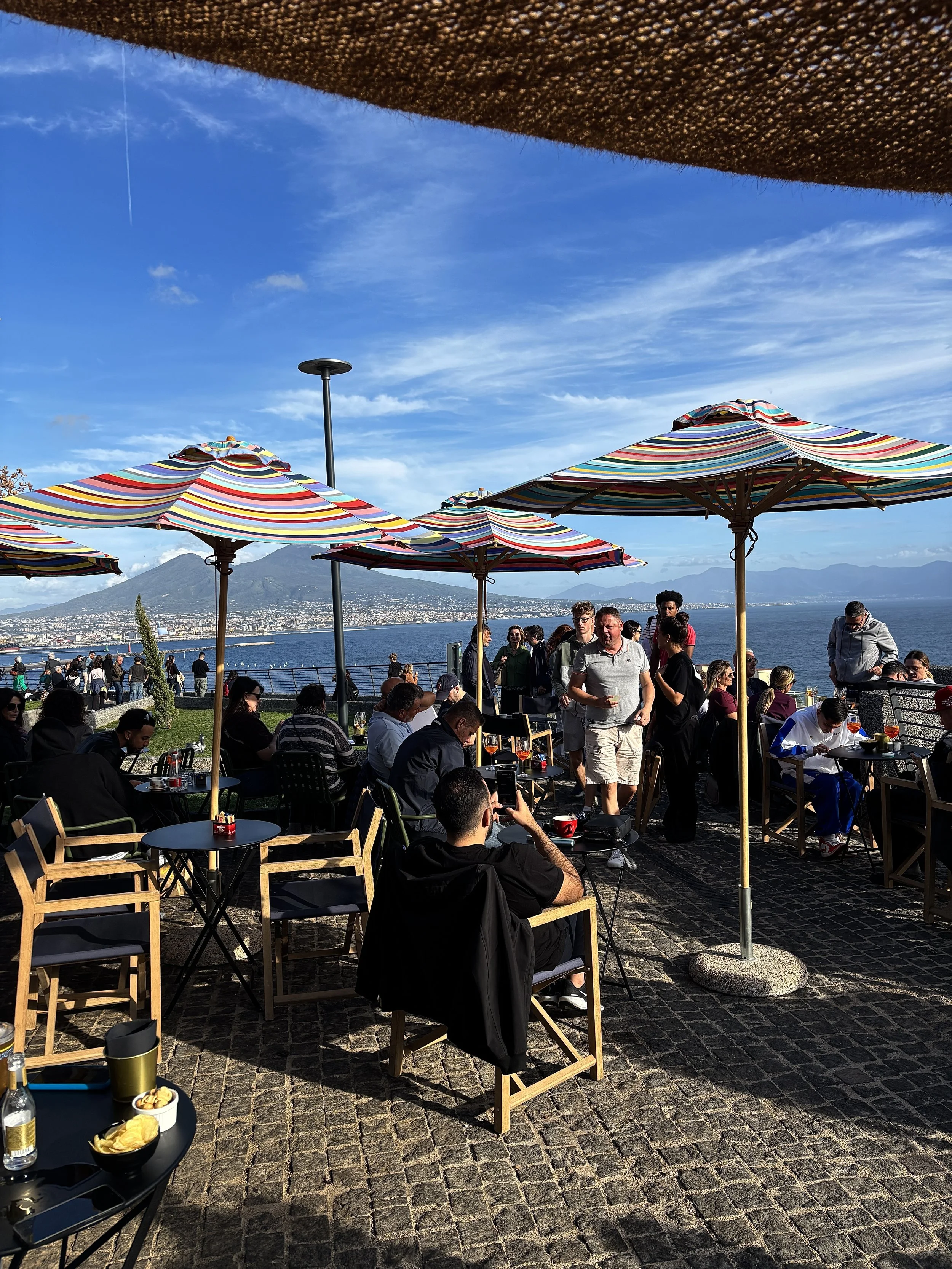 Menschen sitzen und stehen auf einer Terrasse mit bunten Sonnenschirmen, mit Blick auf einen See und Berge im Hintergrund.