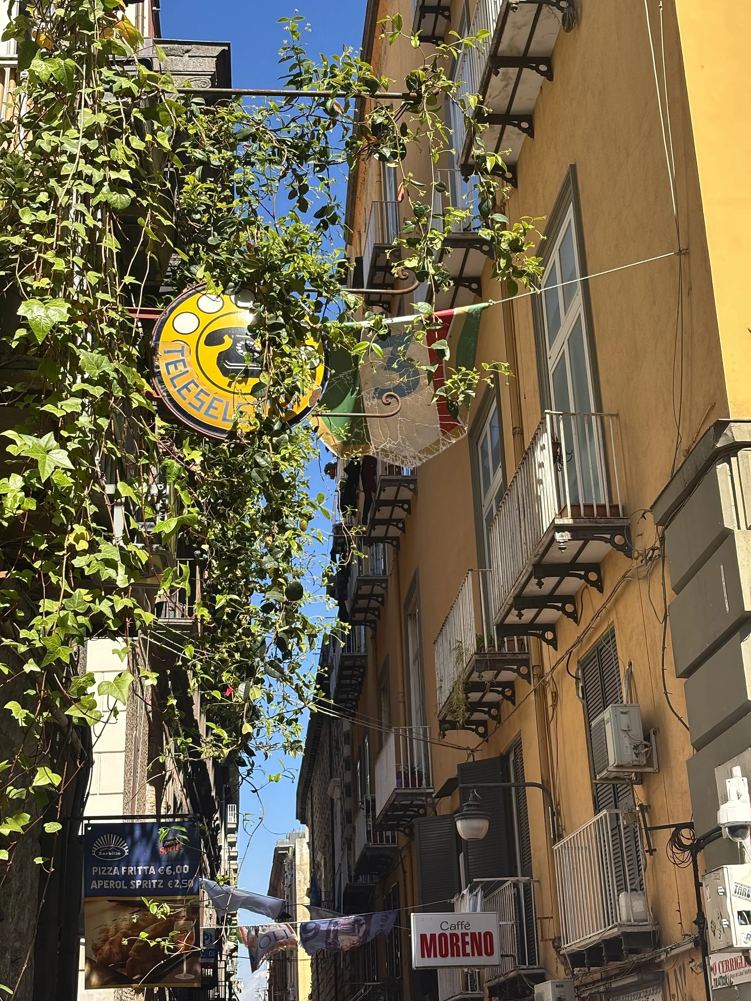 Blick auf eine schmale Straße in Italien mit gelben Gebäuden, mehreren kleinen Balkonen, Straßenlaternen und Geschäften. Es hängen Flaggen und ein Schild für eine Pizzeria. Der Himmel ist blau.