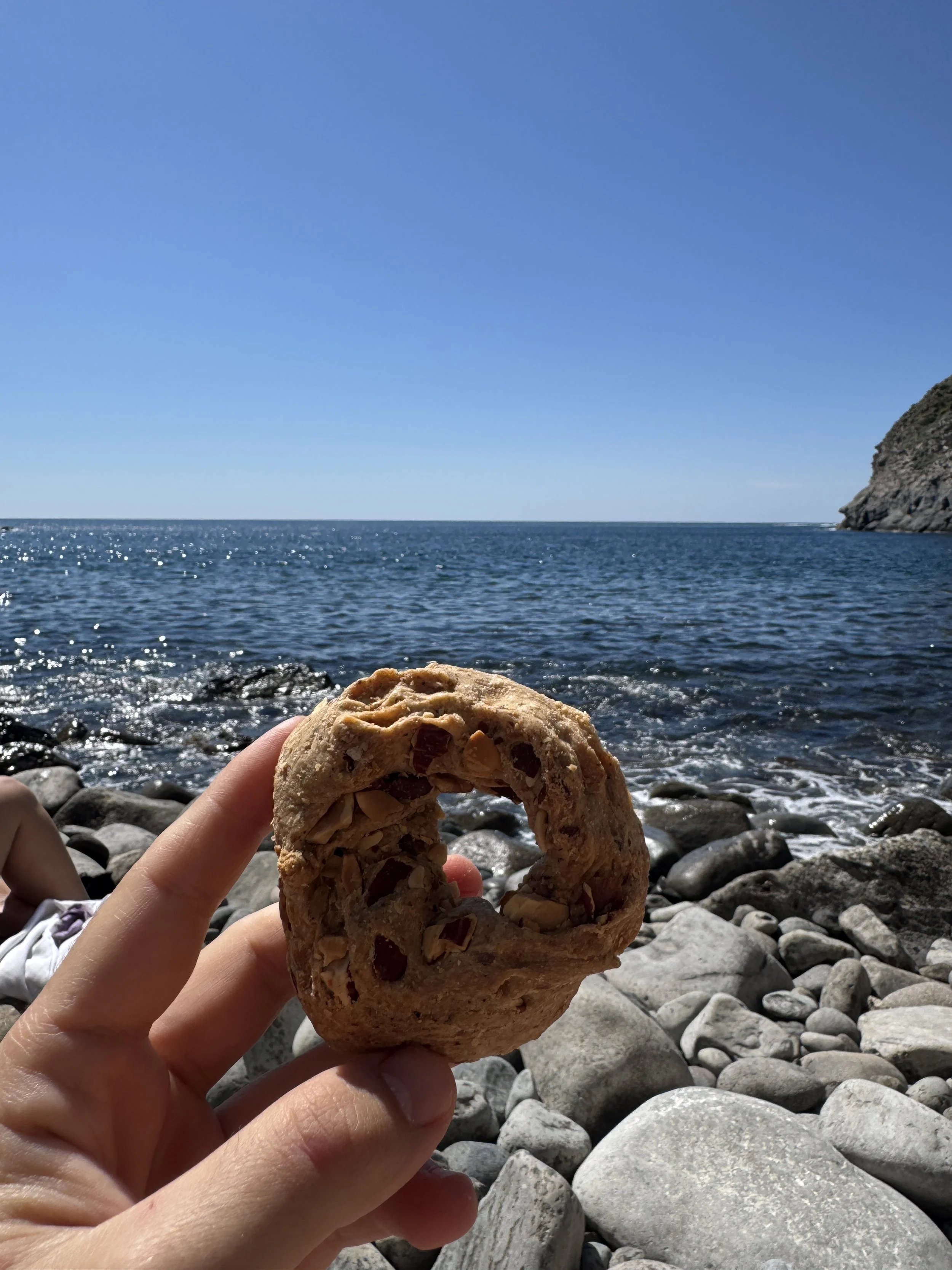 Eine Hand hält einen Keks vor einer Küstenlandschaft mit Felsen, dem Meer und einem klaren blauen Himmel.