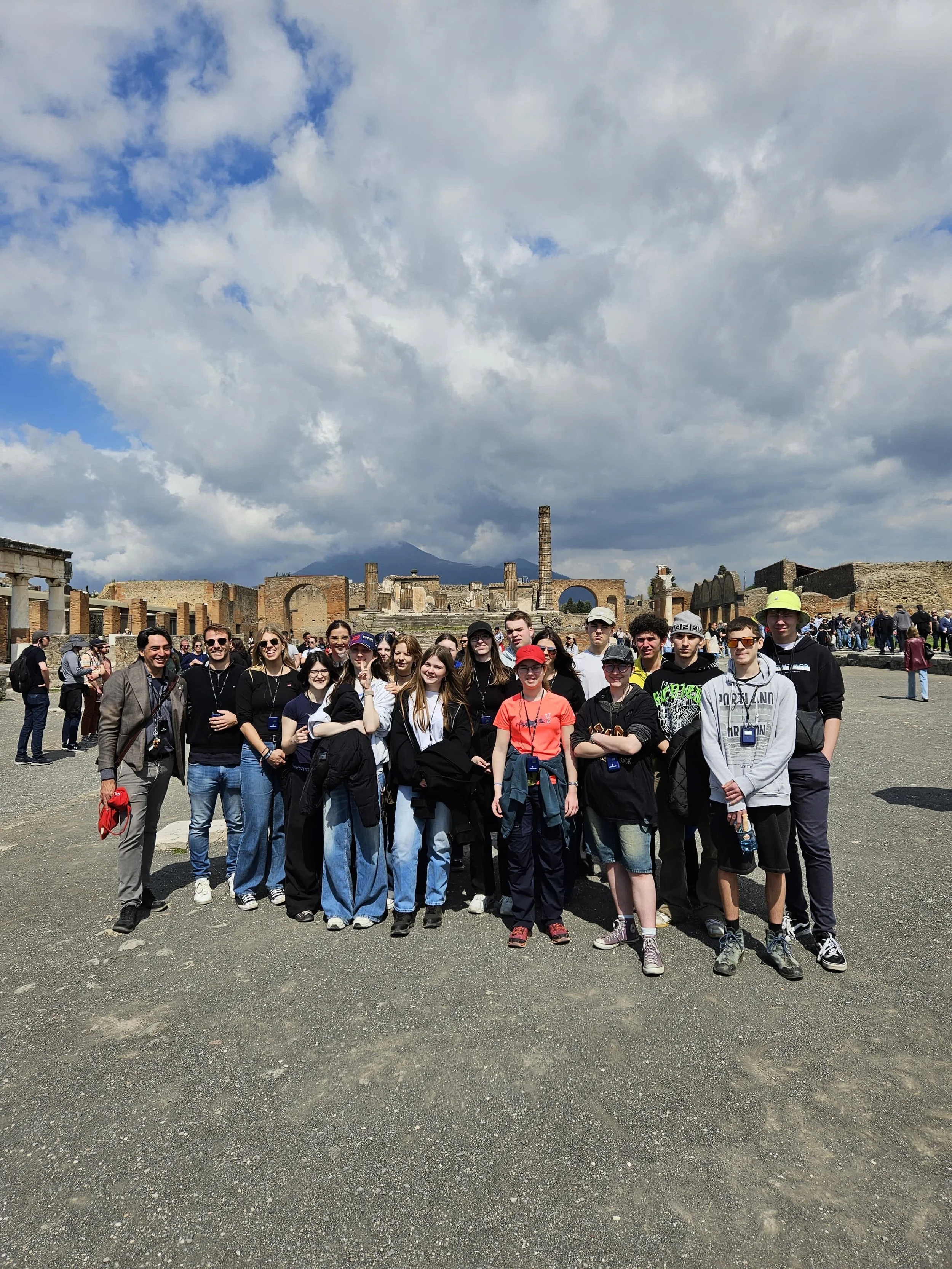 Gruppe von Touristen vor den Überresten antiker Ruinen in Pompeji, mit Bergen im Hintergrund bei bewölktem Himmel.