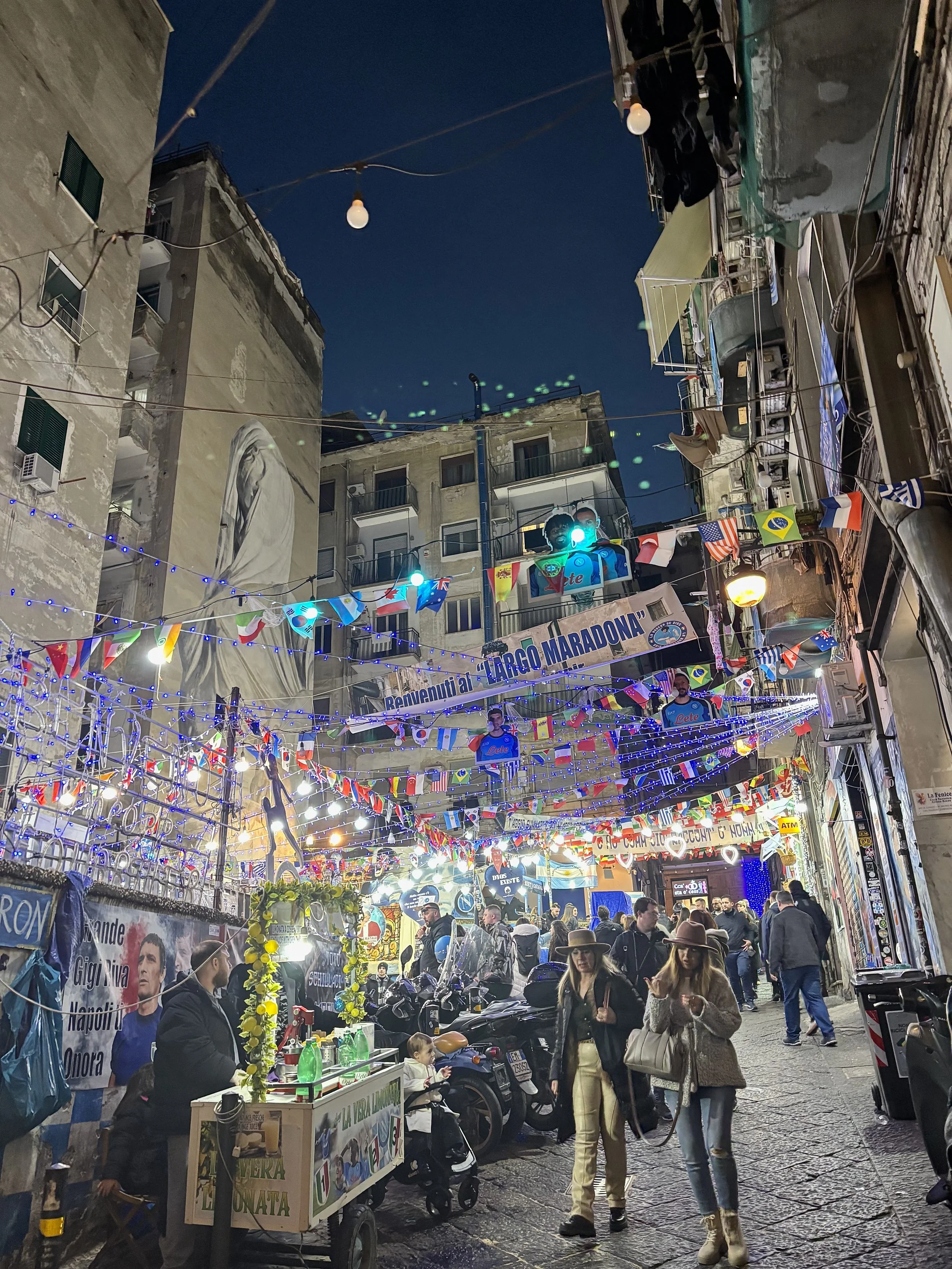 Nachtaufnahme einer belebten Straße in Italien, geschmückt mit Lichterketten und Flaggen, mit Menschen, die spazieren gehen und Geschäfte. Eine Wand mit Graffiti und ein Straßenstand sind sichtbar.