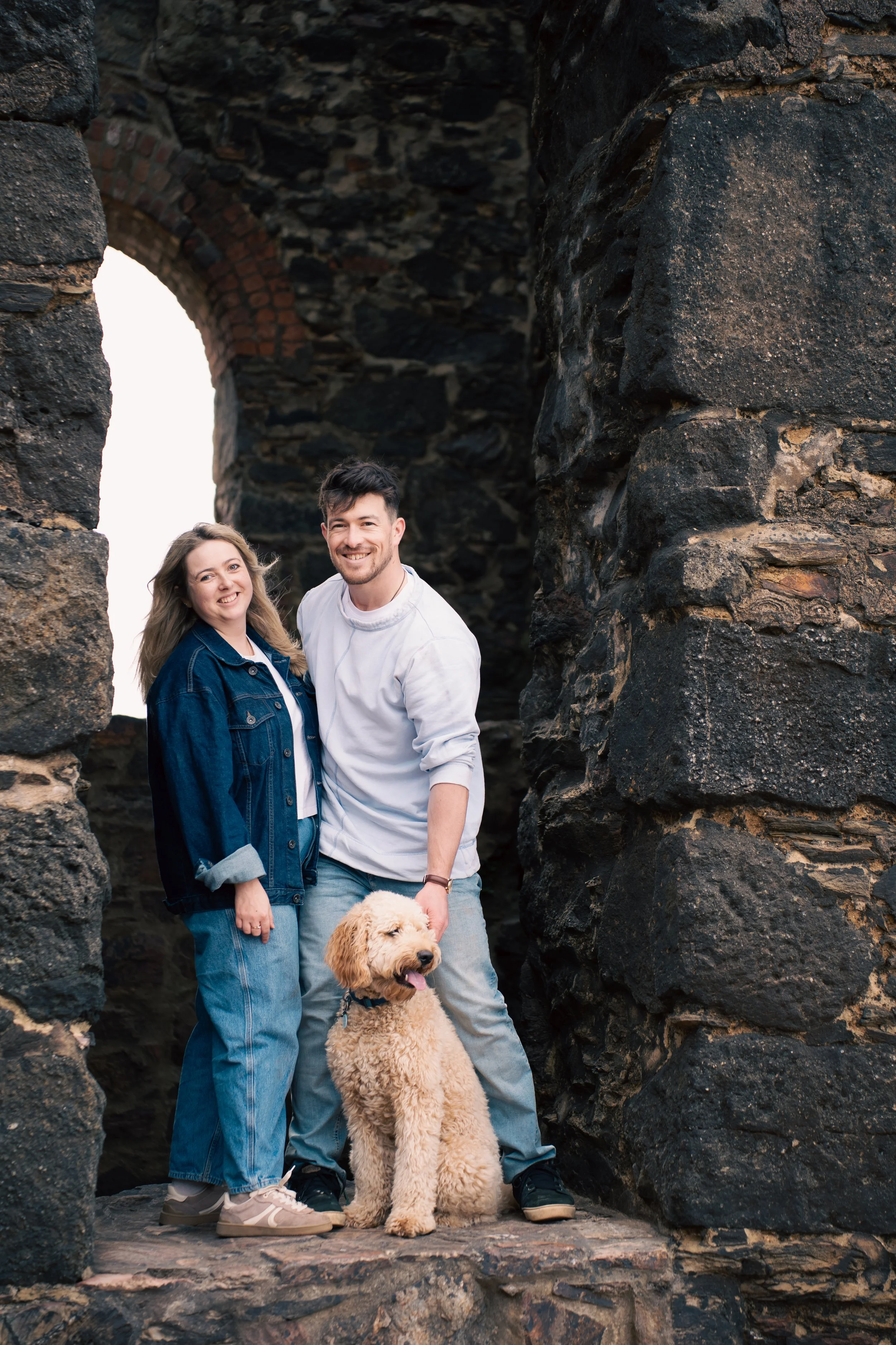 A couple with a golden retriever puppy inside a stone archway. The woman has long, light brown hair and is wearing a denim jacket. The man has short dark hair and is wearing a white sweatshirt. The dog is sitting in front of them, facing forward with its mouth slightly open. The setting appears to be outdoors, with the stone structure showing signs of age and weathering.