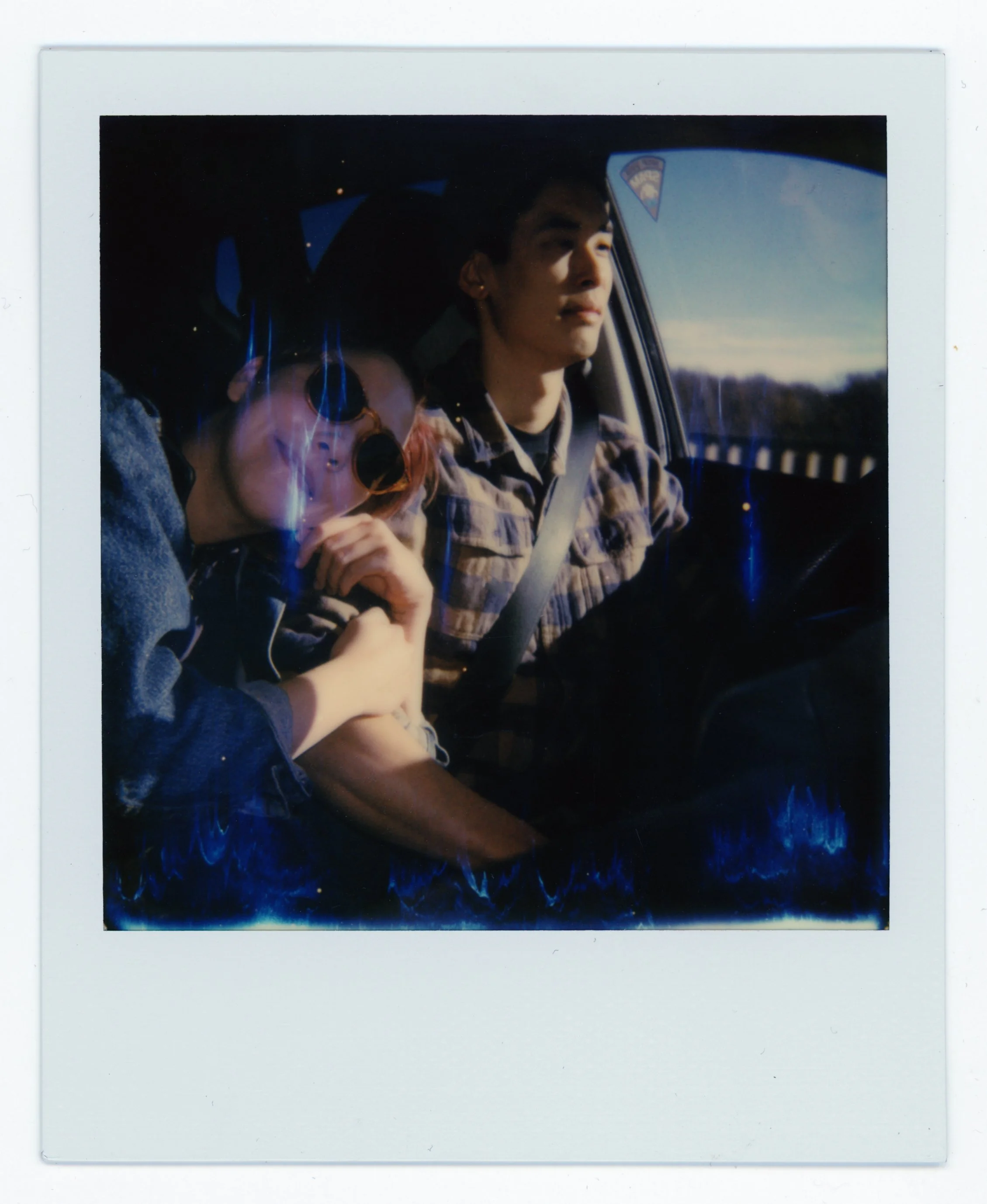 Two people riding in a car, a woman with glasses and earrings leaning on the man's shoulder, during daytime with sunlight and clear skies visible outside.