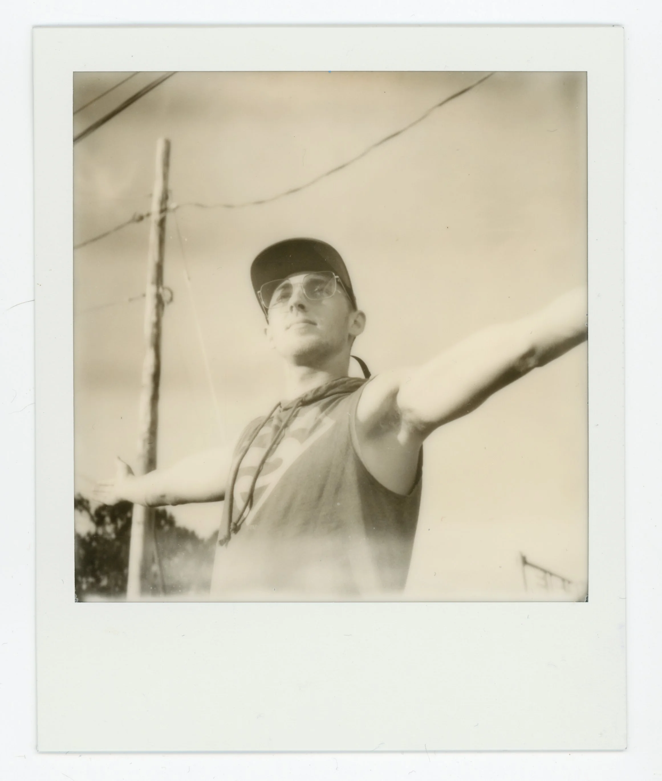A young man taking a selfie with arms outstretched outdoors, wearing a cap, glasses, and a sleeveless shirt, with utility poles and power lines in the background.