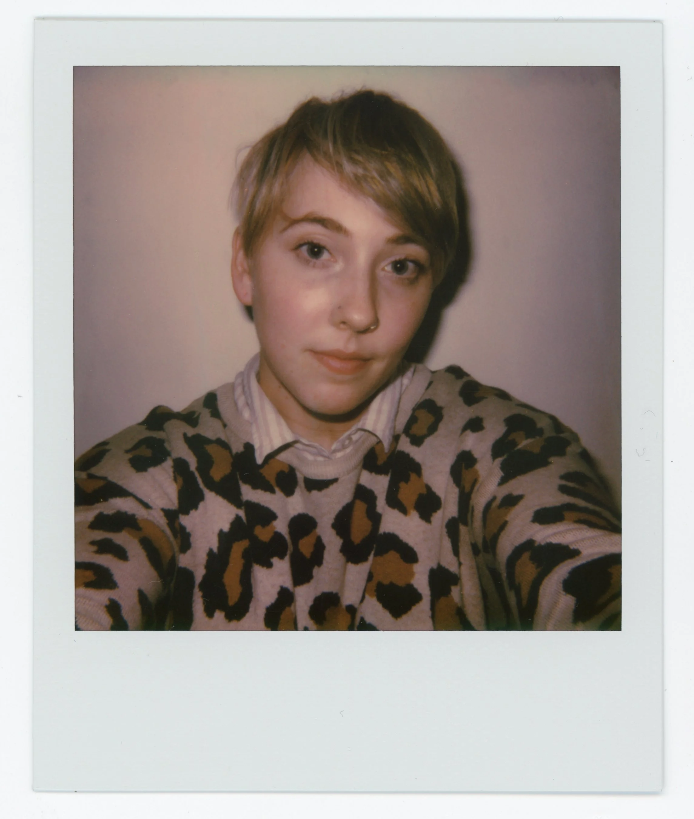 A young woman with short blonde hair wearing a leopard print sweater takes a selfie against a plain background.