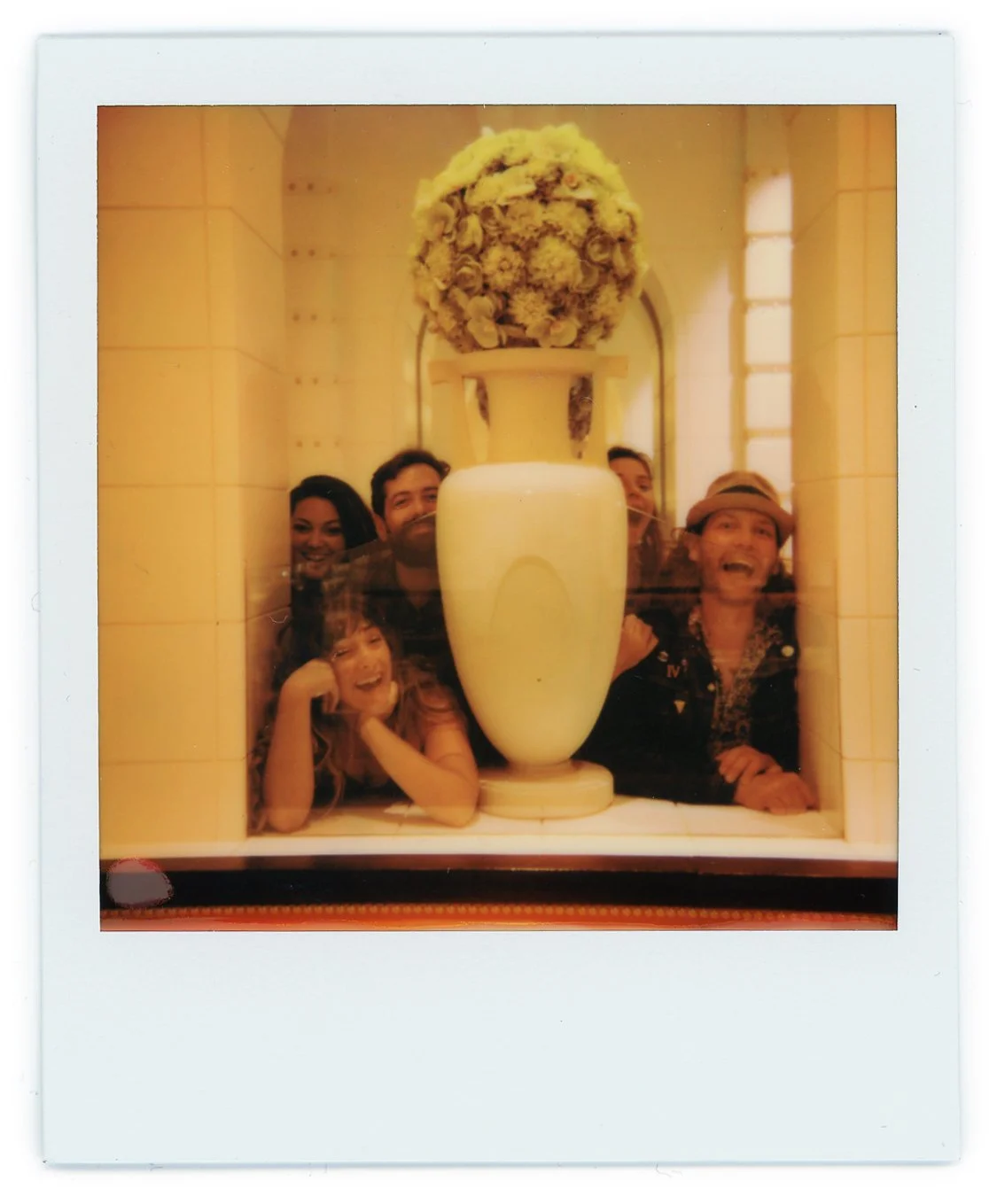 Group of five people behind a large white vase with a flower arrangement on top, smiling and laughing.