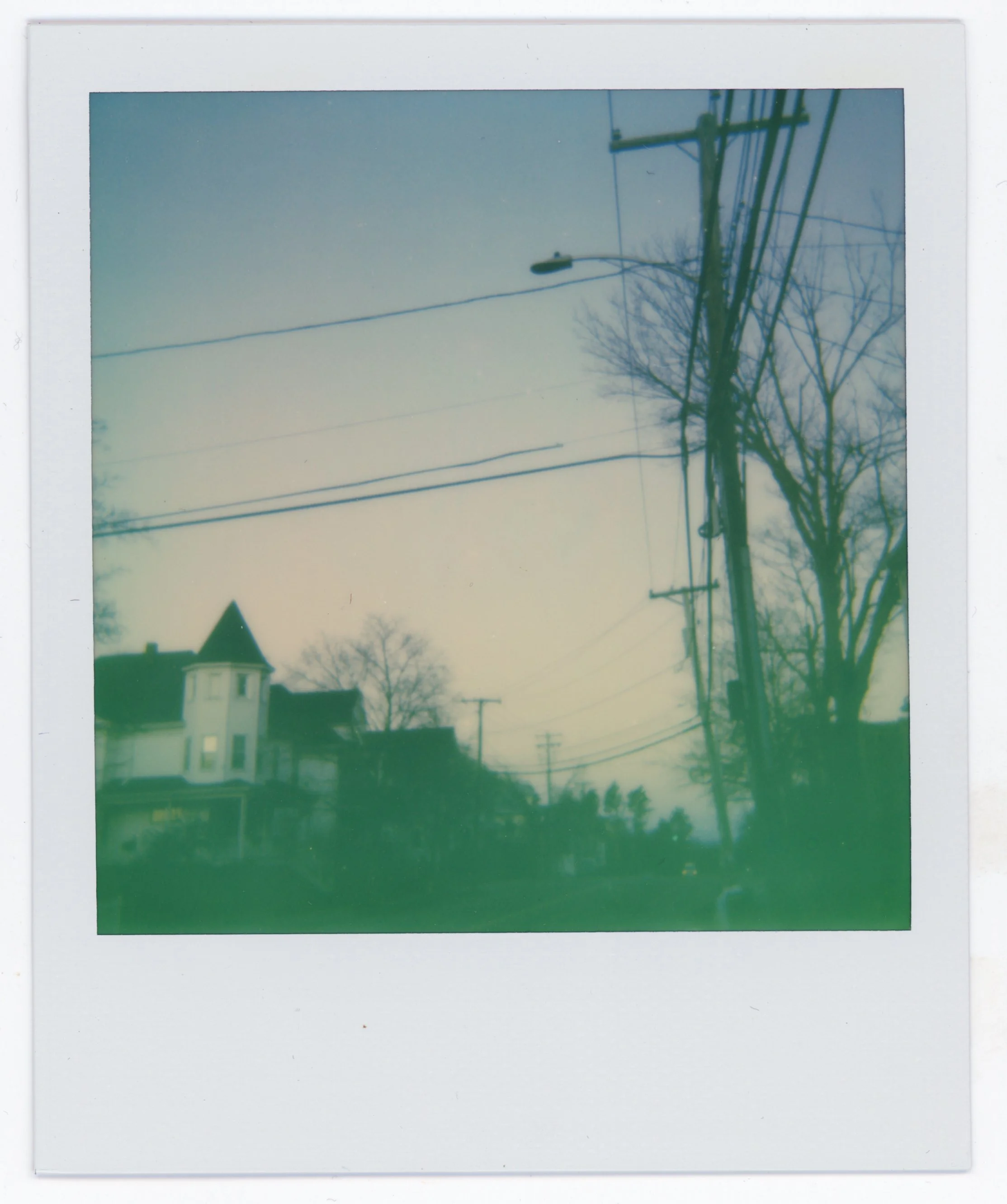 A blurry Polaroid photo of a residential street with utility poles and leafless trees, with a house that has a turreted section on the left.