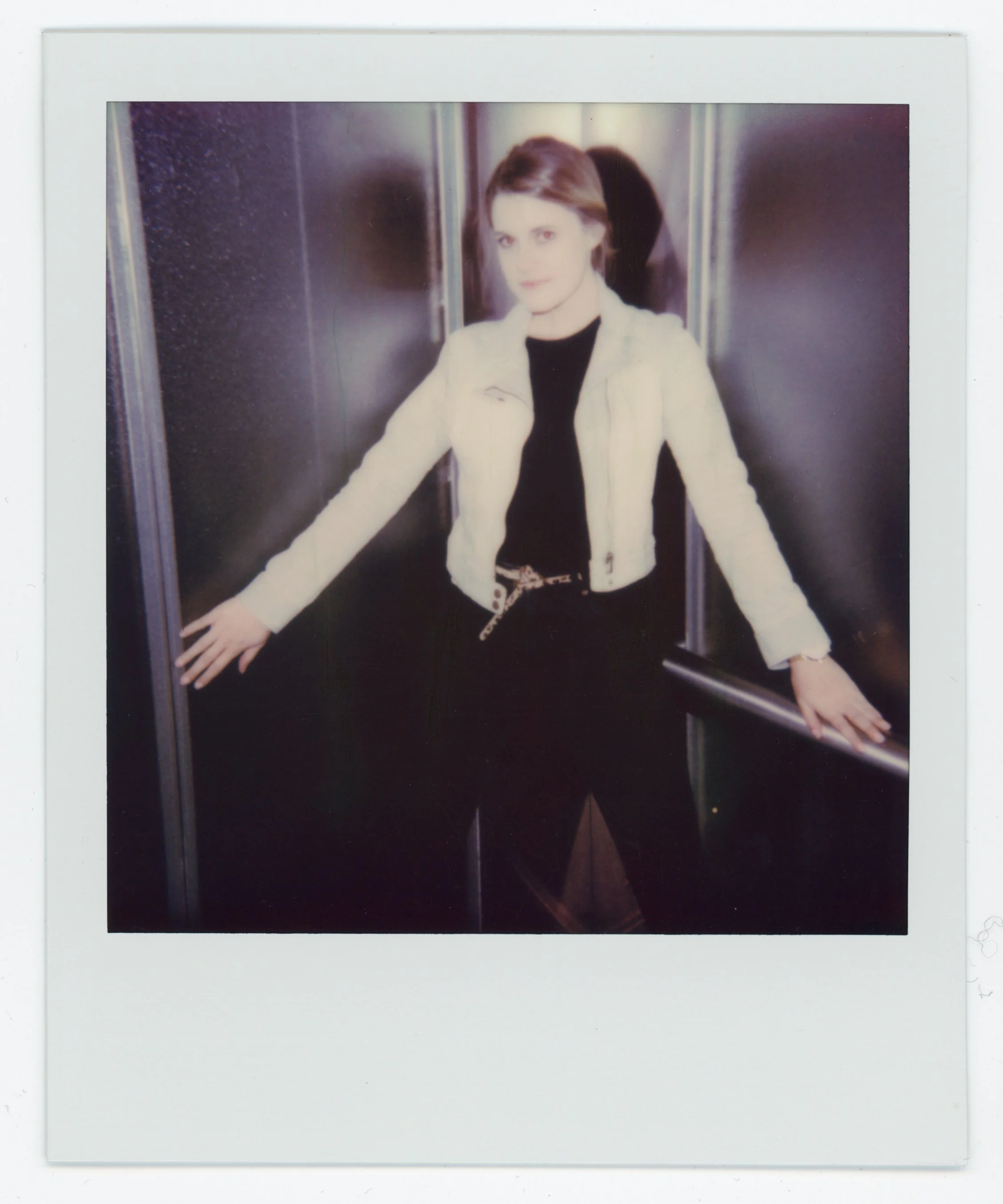 A woman with short brown hair standing in an elevator, leaning on the handrails with her arms outstretched, wearing a light-colored jacket and black clothing.