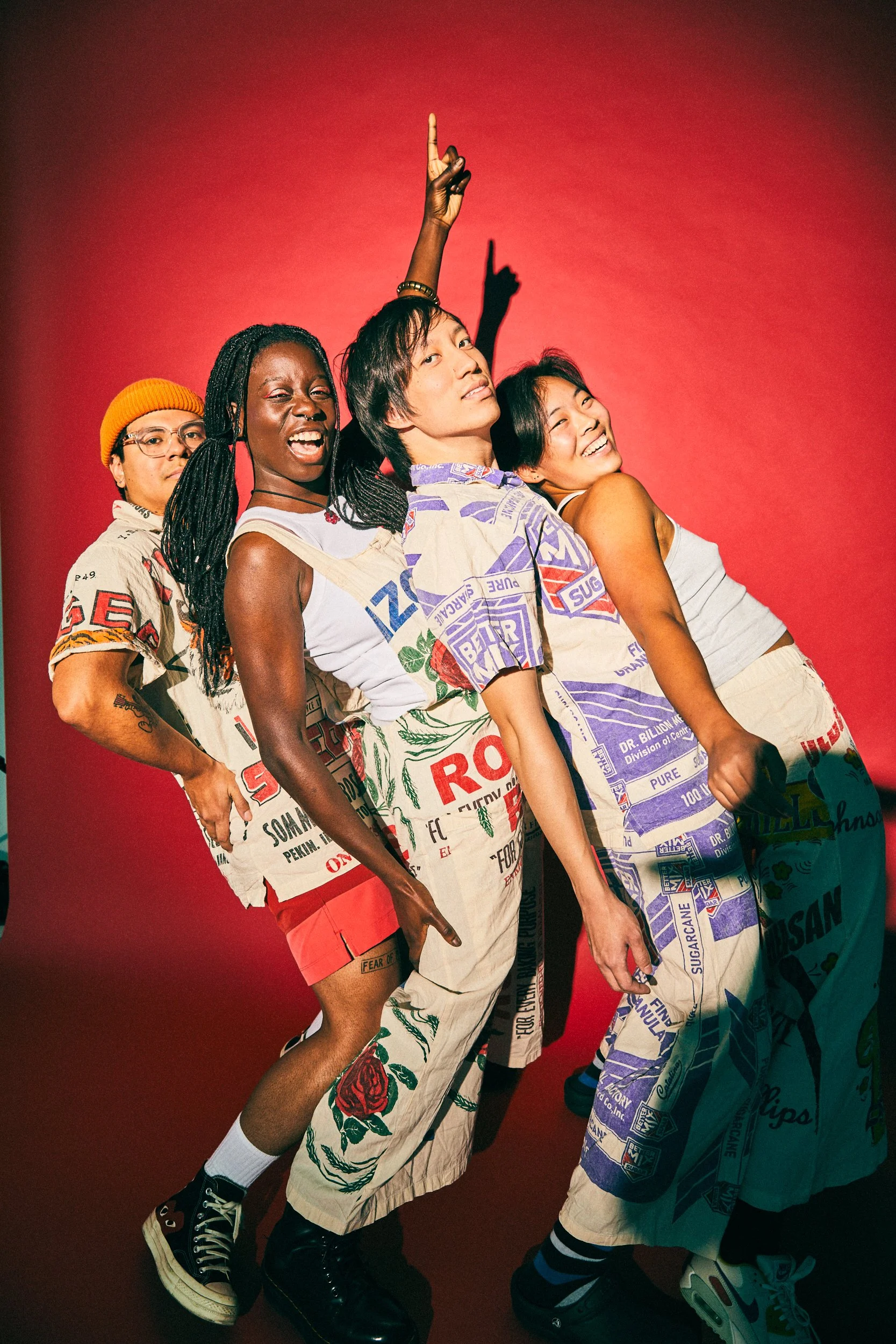 Four diverse young people standing close together against a red background, smiling and posing confidently, with one person pointing upward.