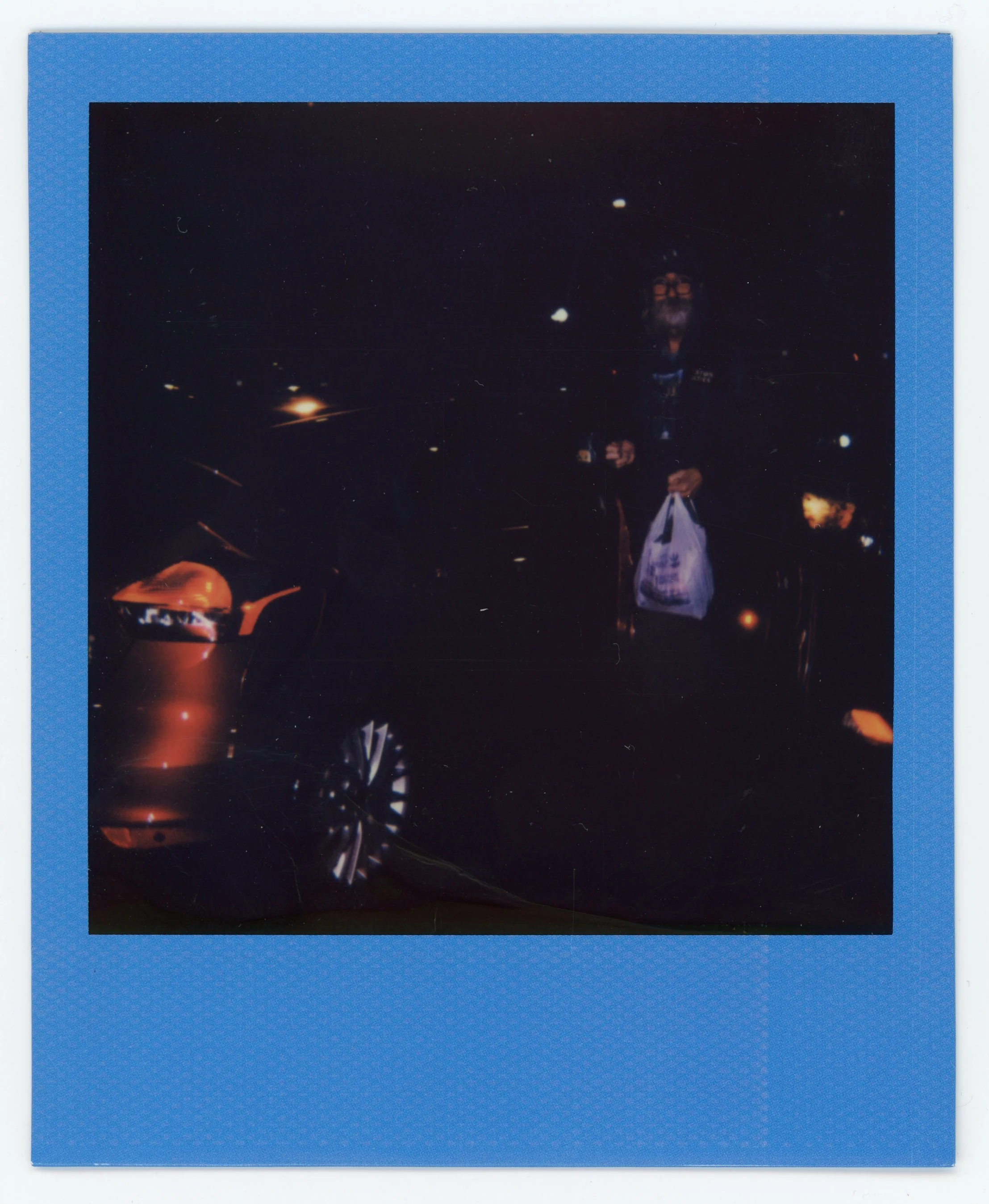 A person standing next to an orange car holding a shopping bag at night, with streetlights and other vehicles in the background.