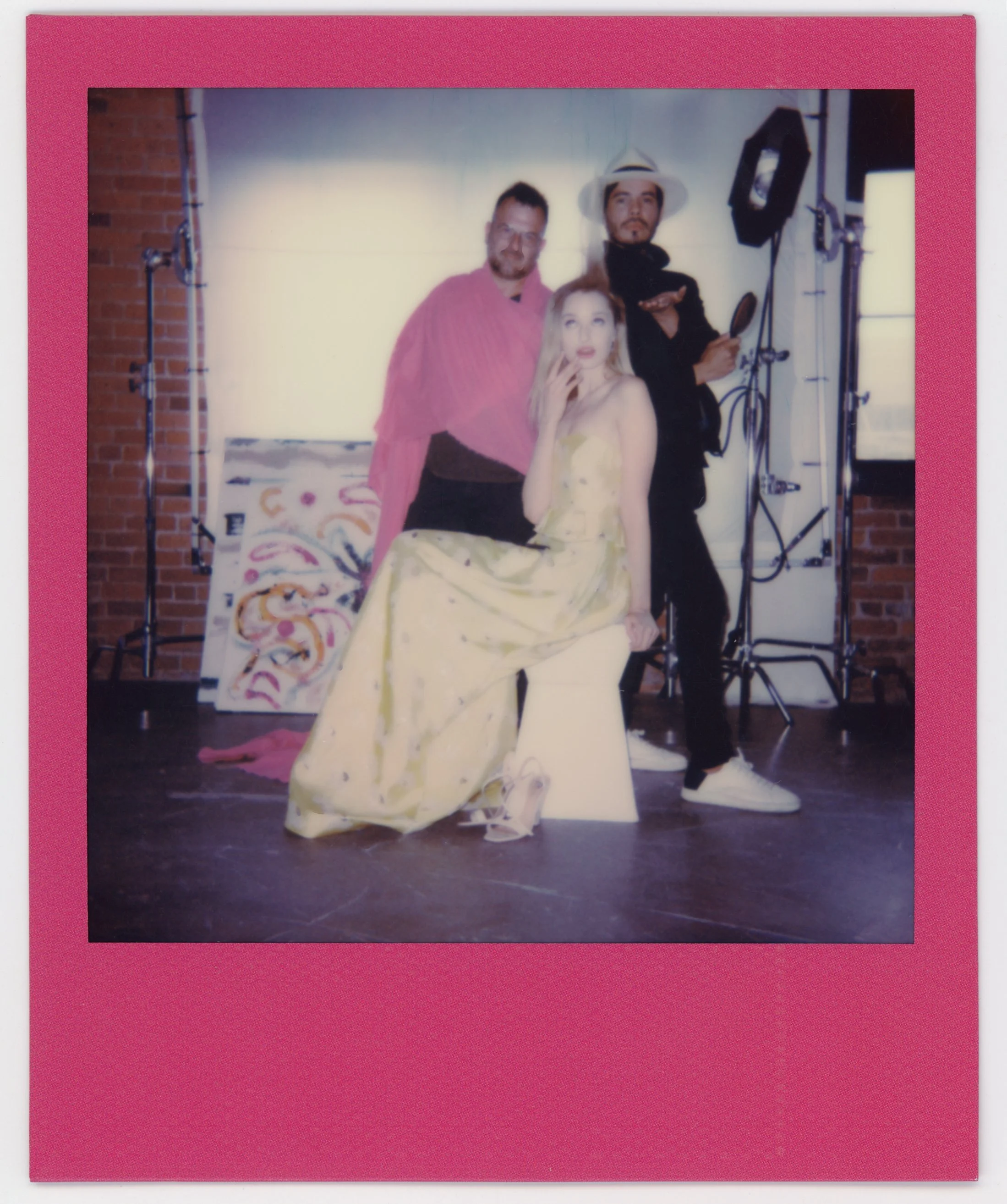 Three people posing in a studio with art supplies and light equipment in the background. One woman in a long, yellow floral dress sitting on a white stool, with a hand touching her chin. Two men standing behind her, one wearing a pink outfit and the 