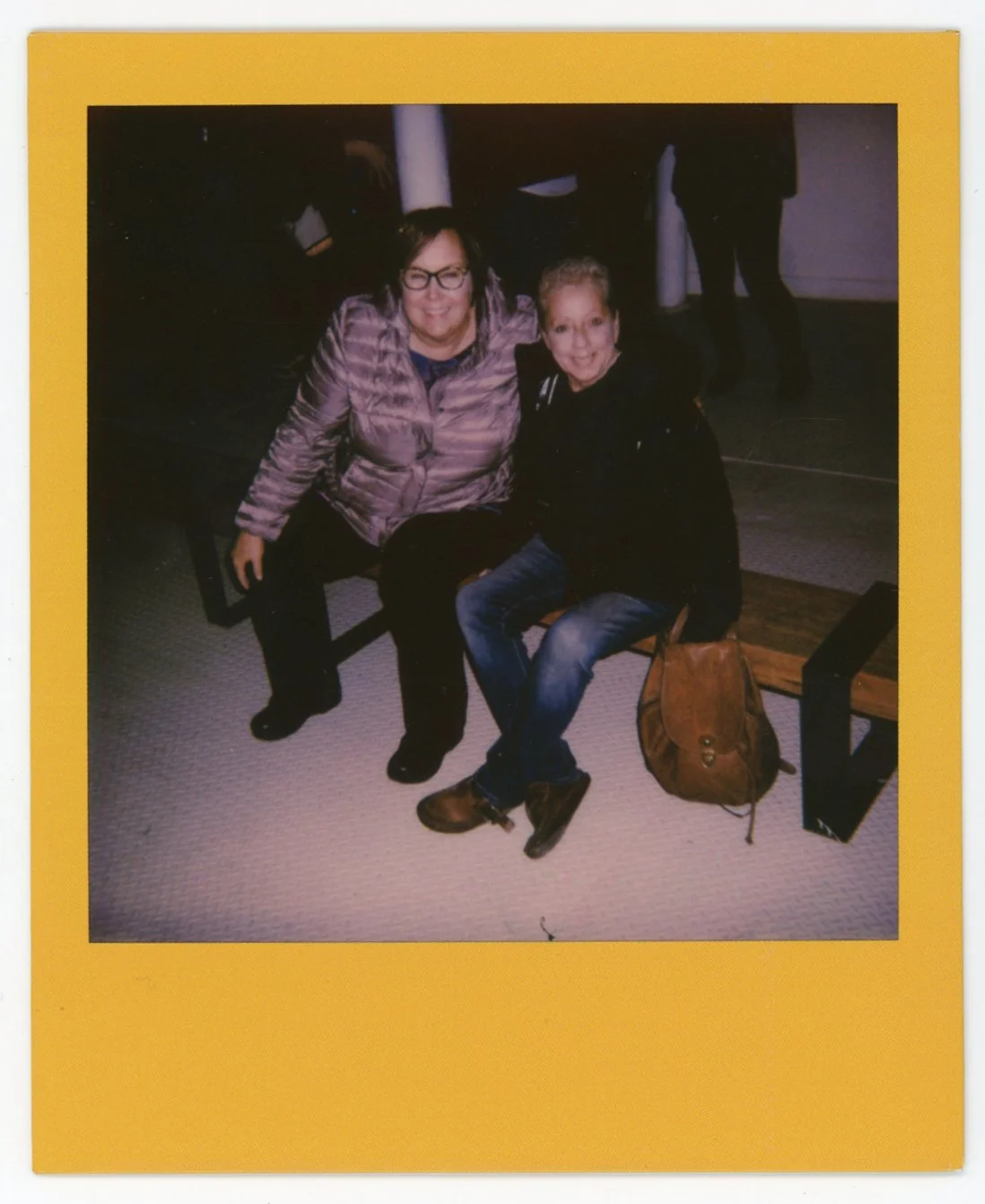 Two women sitting on benches smiling, one in a pink jacket and the other in a black jacket, in an indoor setting with a yellow border.