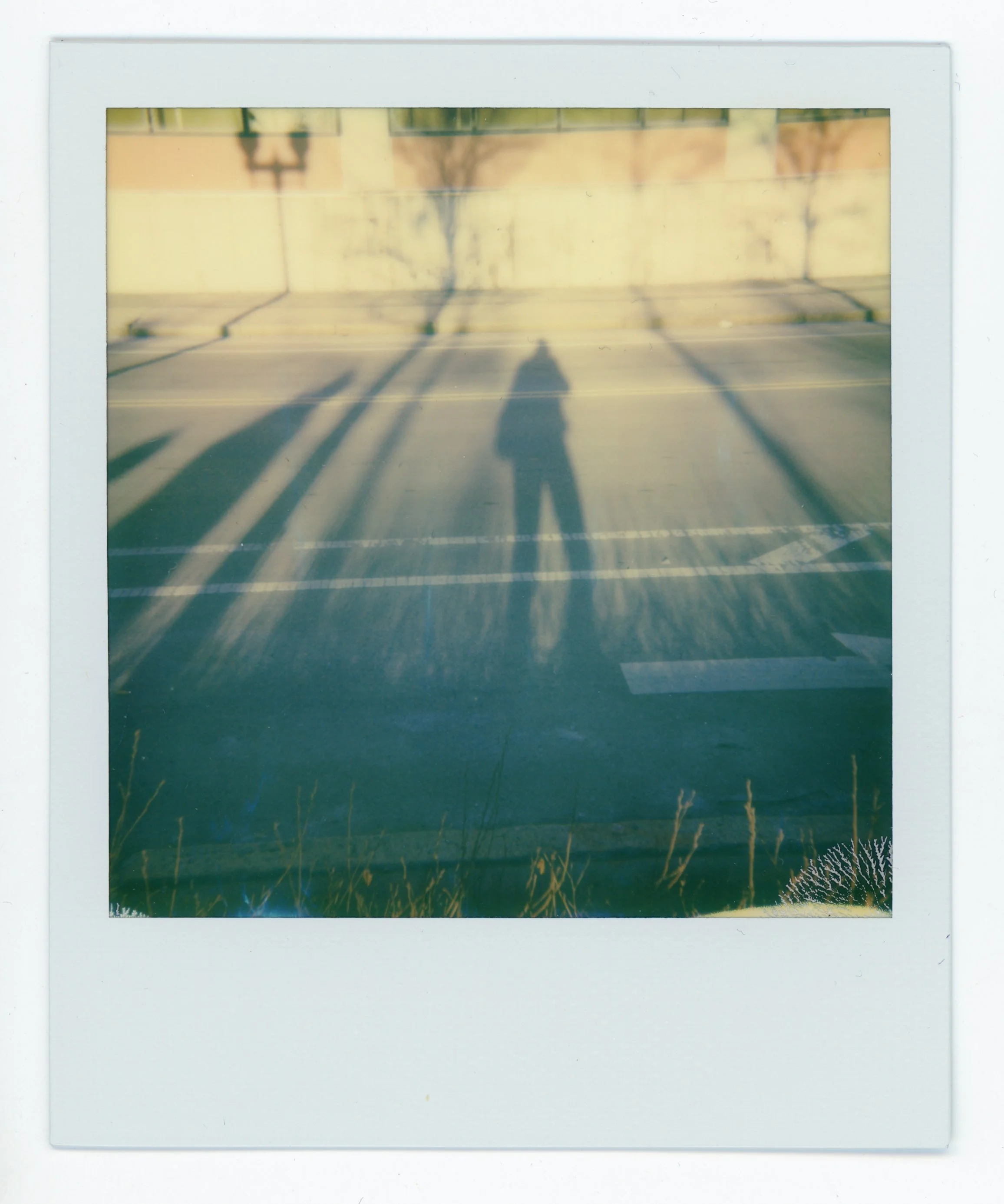 Polaroid photo showing a person's shadow cast on an empty parking lot with three large shadows of streetlights and a faint silhouette of a tree in the background, during sunset.