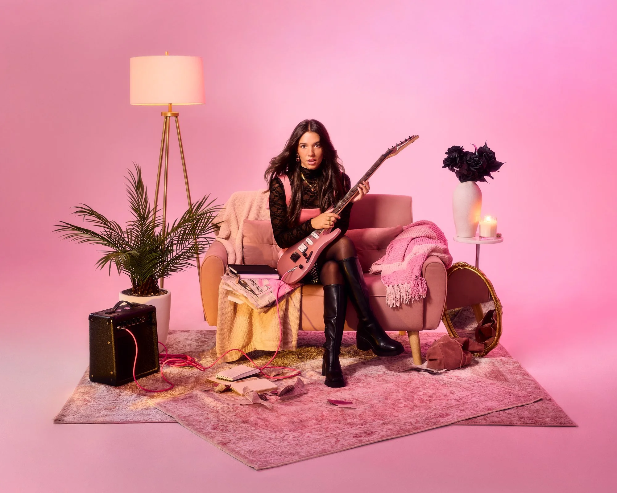 Woman sitting on pink sofa holding an electric guitar, surrounded by pink and black decor, with a pink background, a lamp, a potted plant, and various objects including a small amplifier and candles.