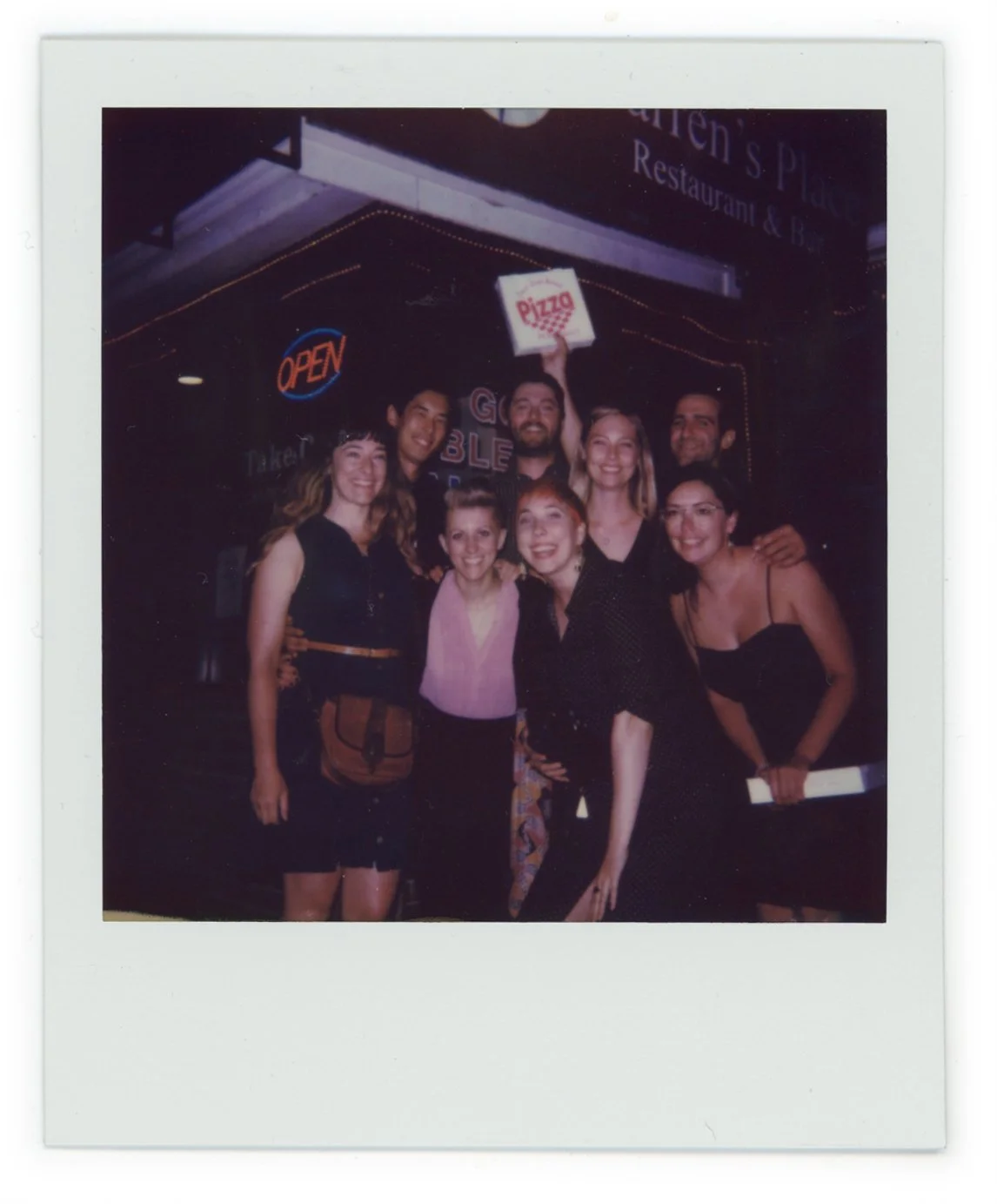 A group of nine people smiling and posing for a photo in front of a neon-lit sign at night. One person is holding a sign that says 'pizza'.