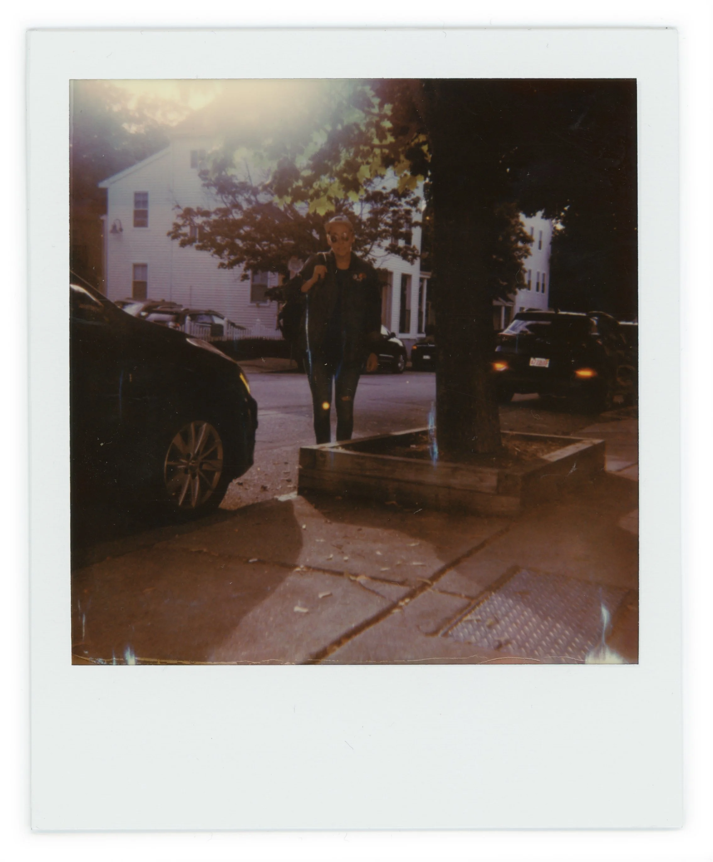Person standing on a sidewalk near parked cars and a tree, with houses in the background and sunlight shining from the top left corner.