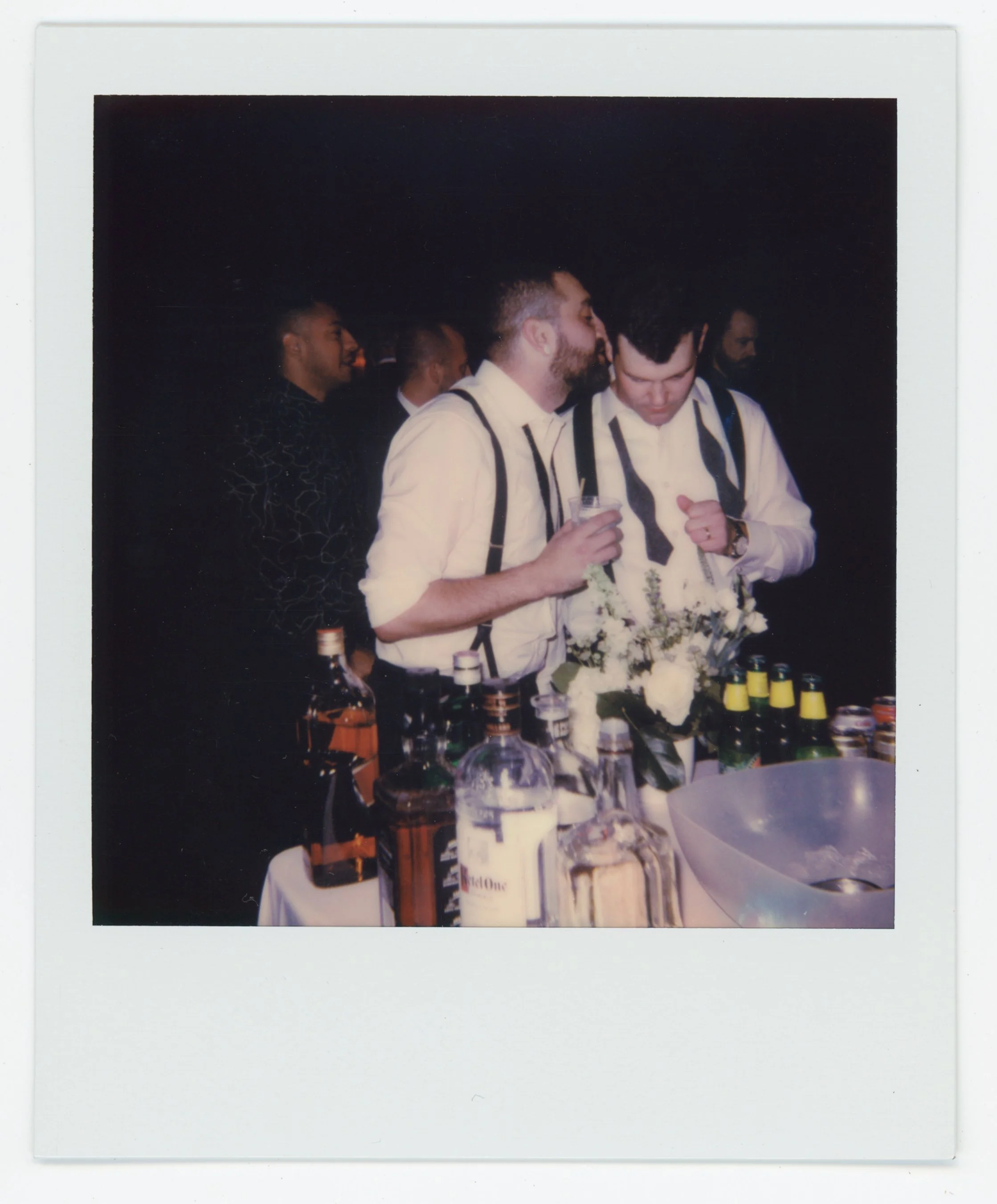 Two men in dress shirts with suspenders standing at a bar or table with bottles and glasses, engaged in conversation, at a social gathering or party.