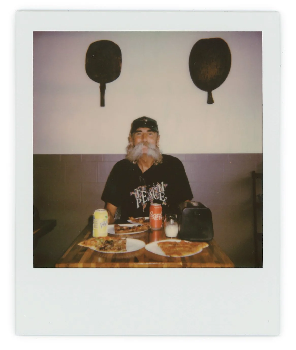 A man with a gray beard and long hair sitting at a table with pizza, soda, and condiments in a restaurant.