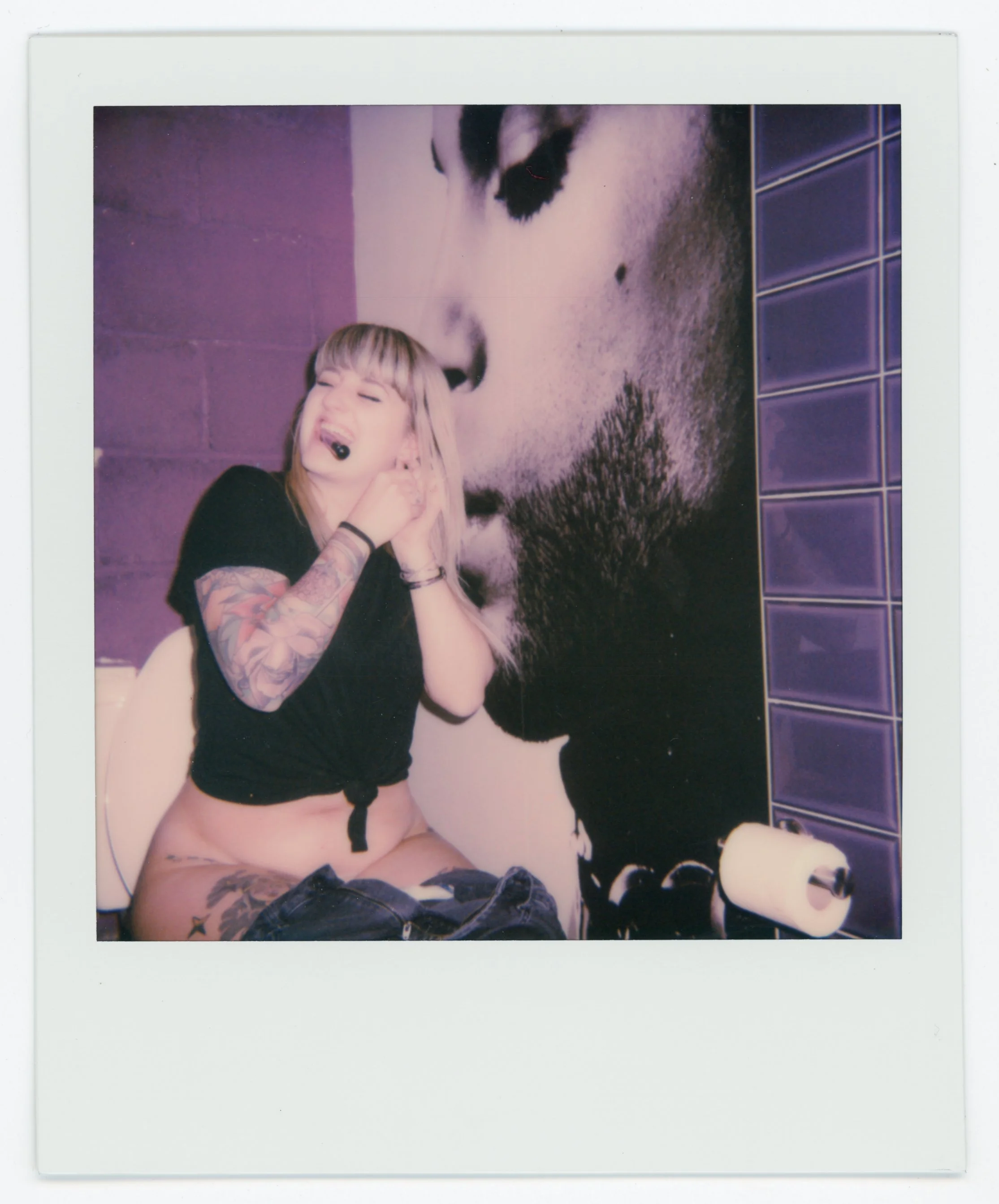 A woman with tattoos laughing and covering her ears, seated next to a large mural of a person's face with purple tiled wall on the right.