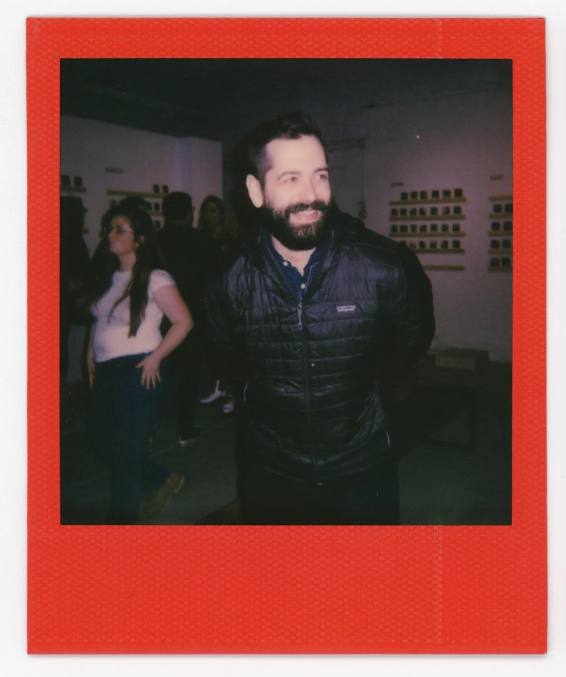 A smiling man with a beard and dark hair, wearing a black quilted jacket, standing in an indoor setting with other people in the background, including a woman with glasses and long dark hair.