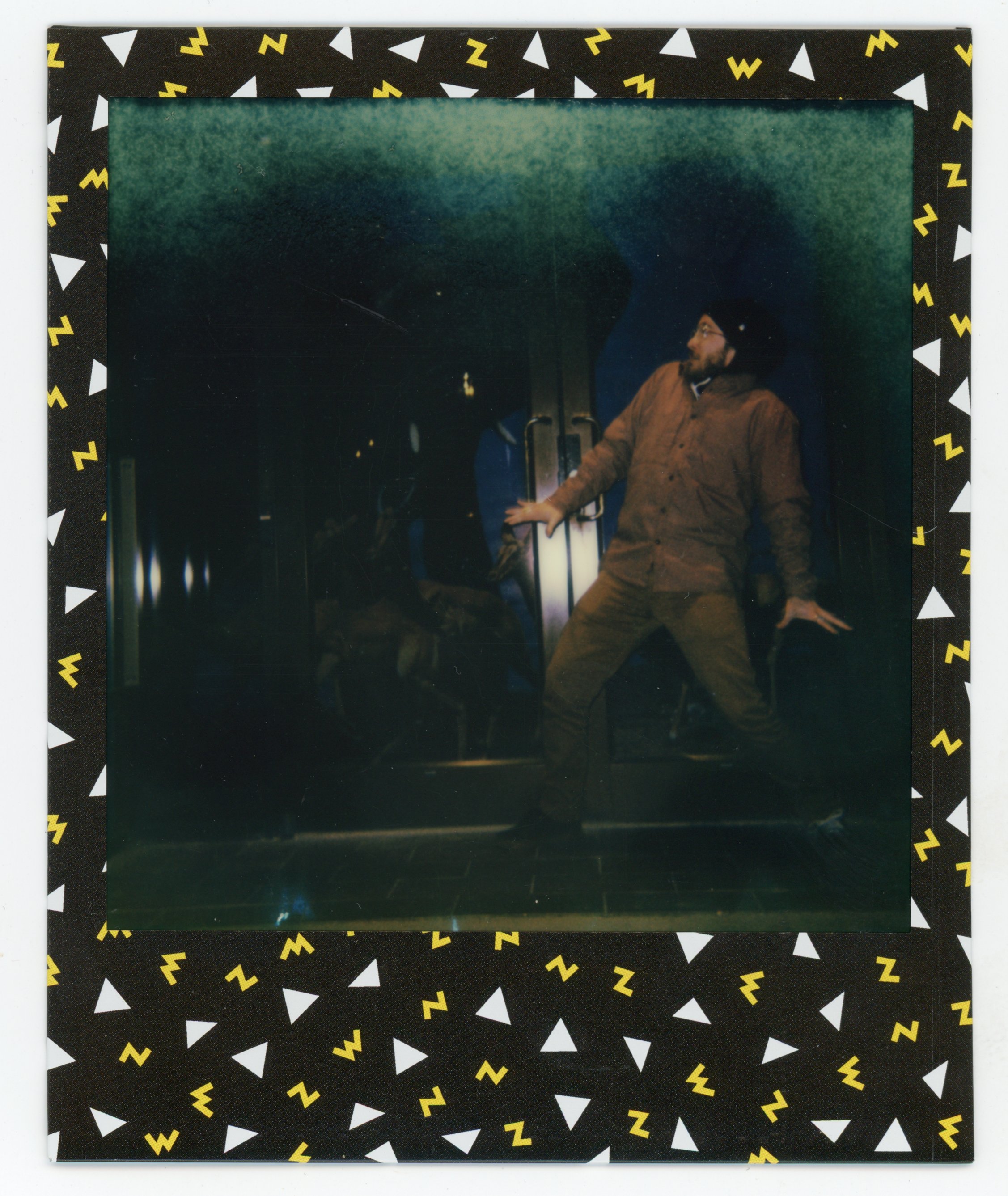 A person wearing a brown jacket and black hat, standing on a dimly lit stage, posing with hands against a door handle, in front of dark background and blue-toned lighting.