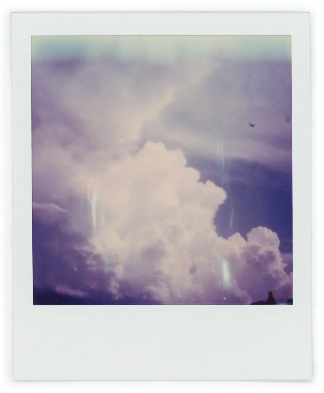Photograph of a cloudy sky with some rainbow-like light refractions and an airplane flying in the distance.