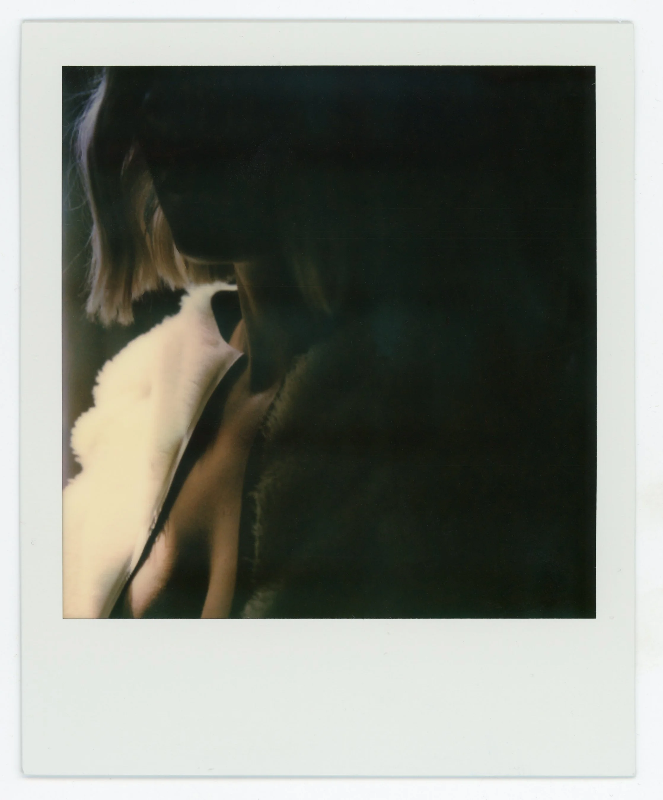 A Polaroid photo of a woman with blonde hair in a black shirt, sitting in a dark room, holding a plush white stuffed animal.