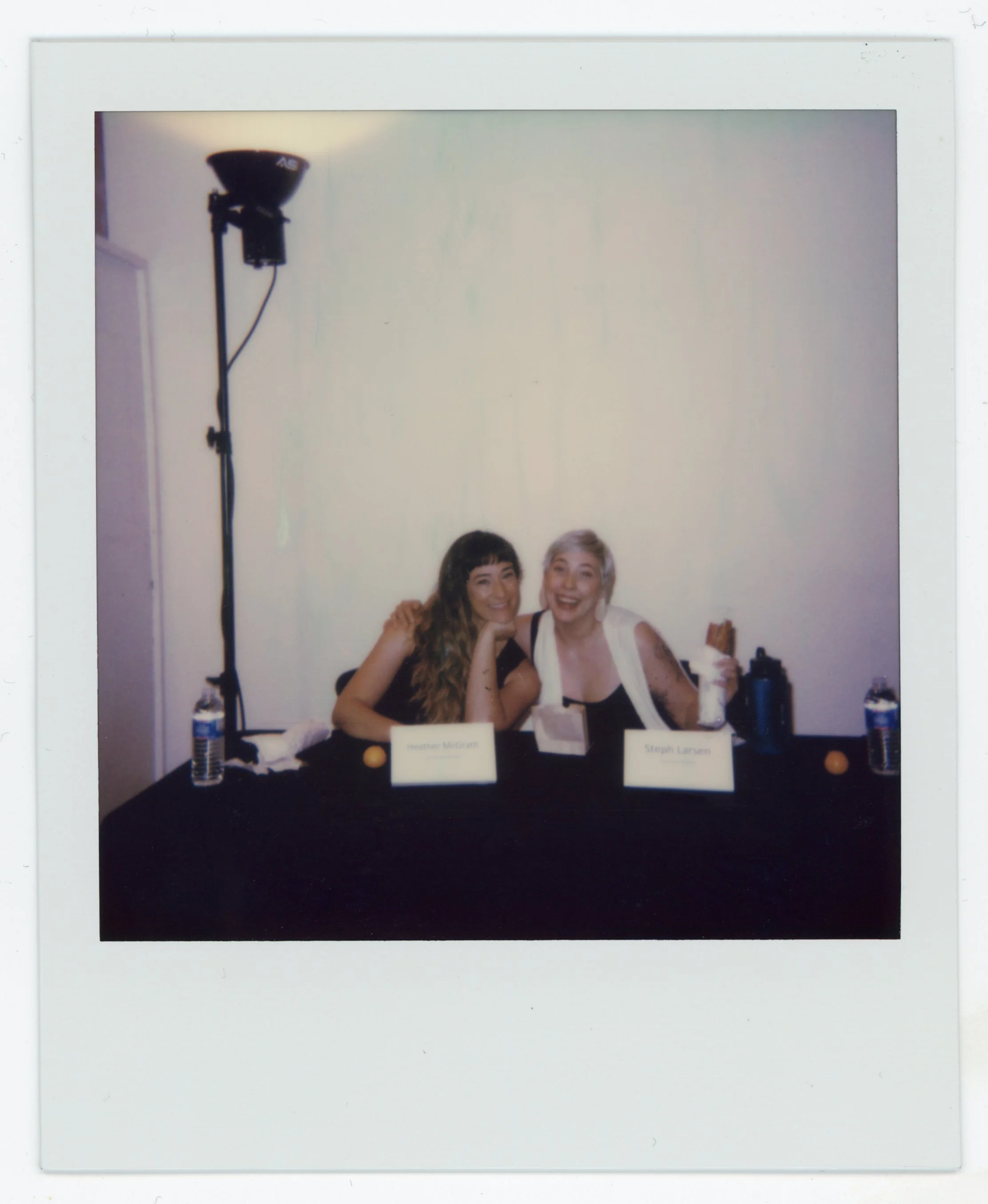 Two women sitting at a table with name tags, water bottles, and snacks, smiling at the camera, with a large light in the background.