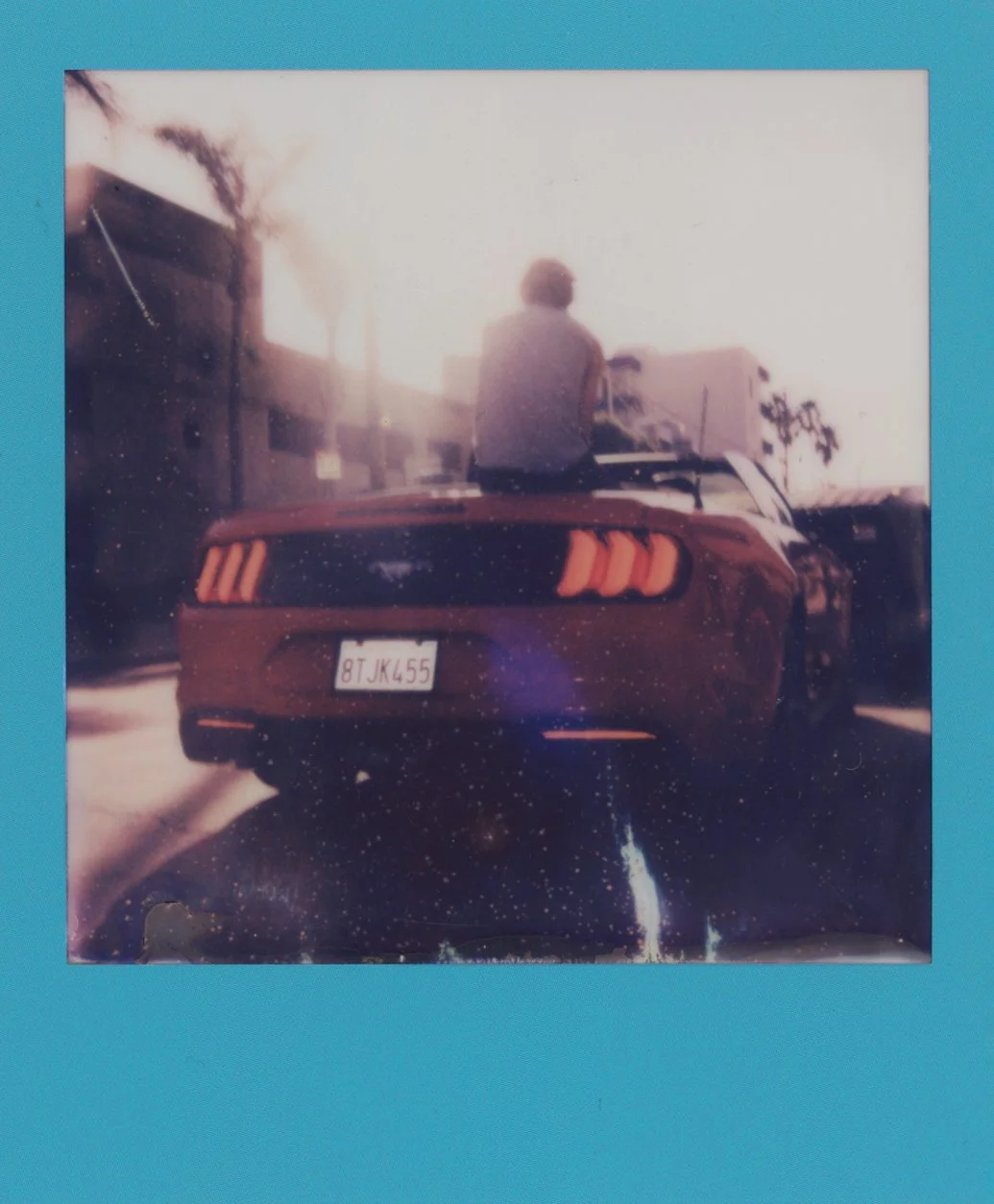 A person sitting on top of a red Ford Mustang car, facing away, with a city street and palm trees in the background. The photo appears to be old or vintage.