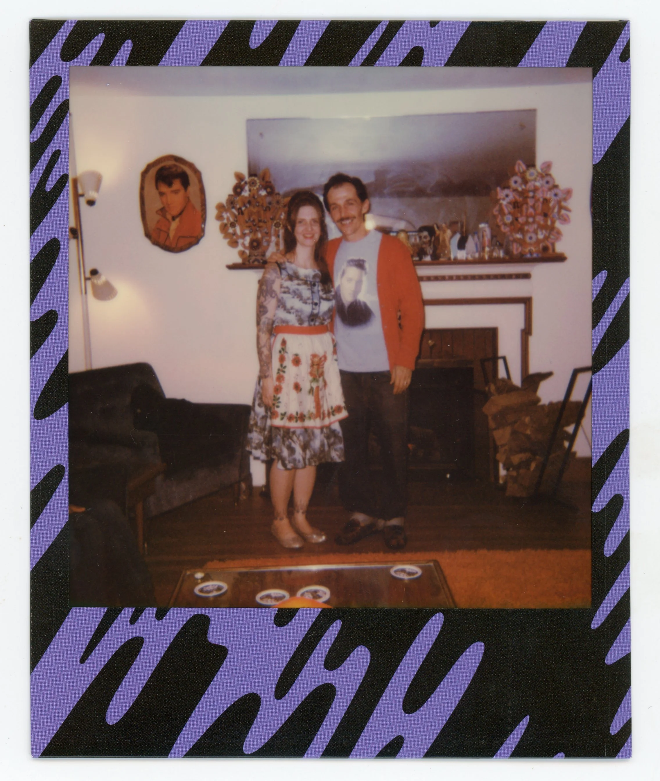 A man and woman standing together in a living room, smiling. The woman wears a floral dress with a red and white apron, and the man wears a graphic t-shirt and an orange cardigan. They are standing in front of a fireplace with decorations and a large