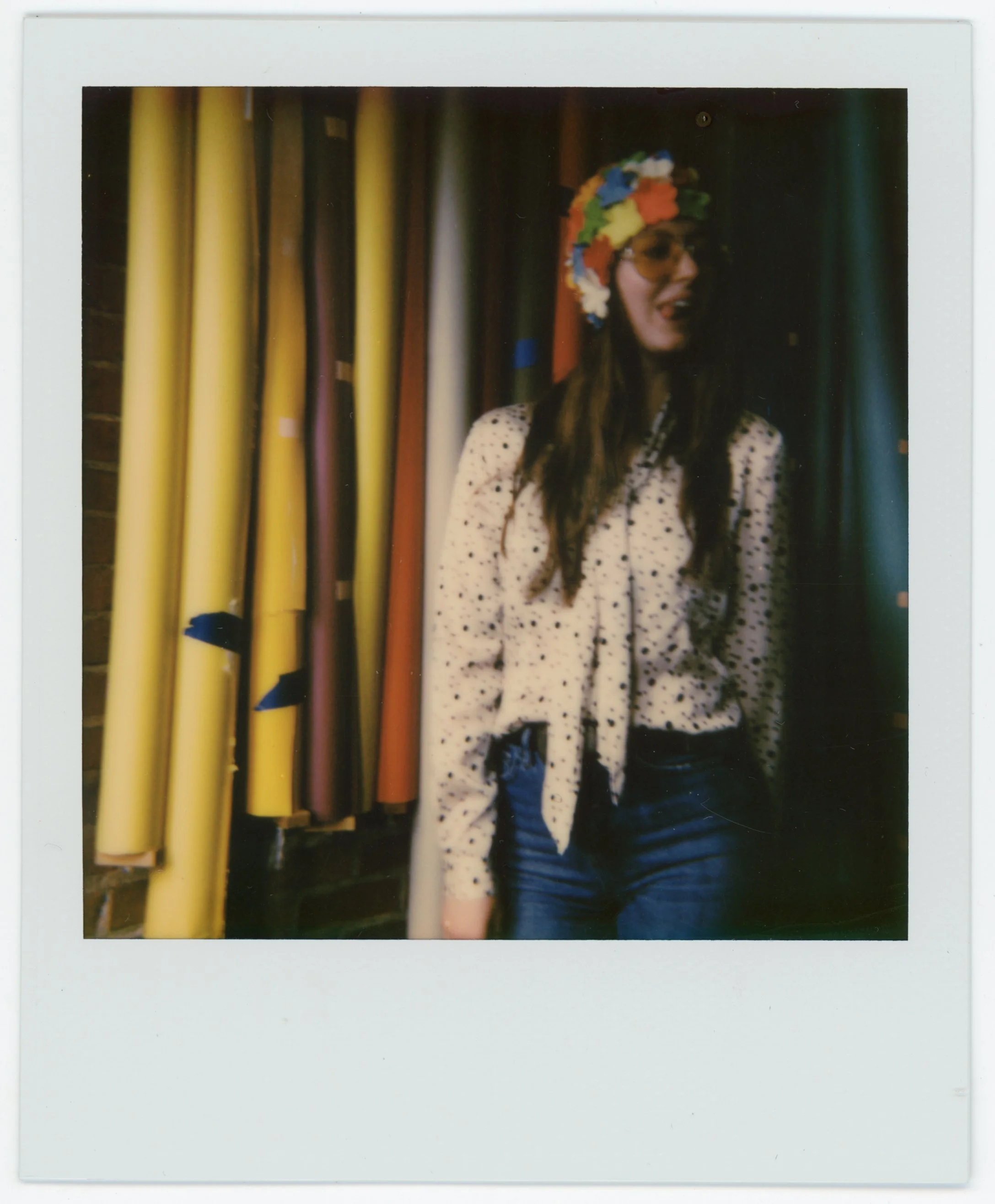 A woman wearing a colorful flower crown and sunglasses, smiling, standing next to fabric rolls in a store or boutique.