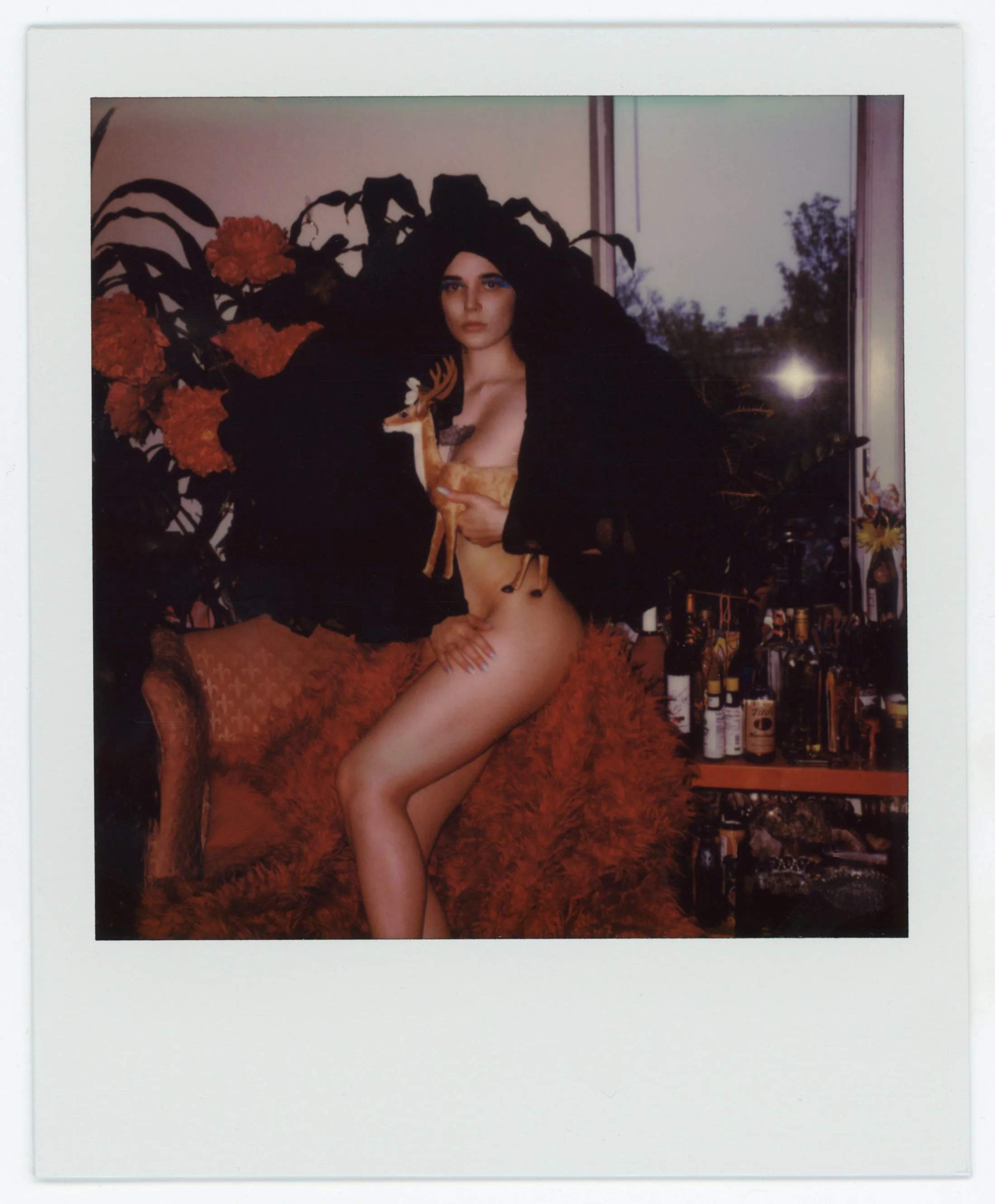 A woman with dark hair and blue eyes standing next to a large floral arrangement, holding a small deer figurine, with a bar and bottles in the background.