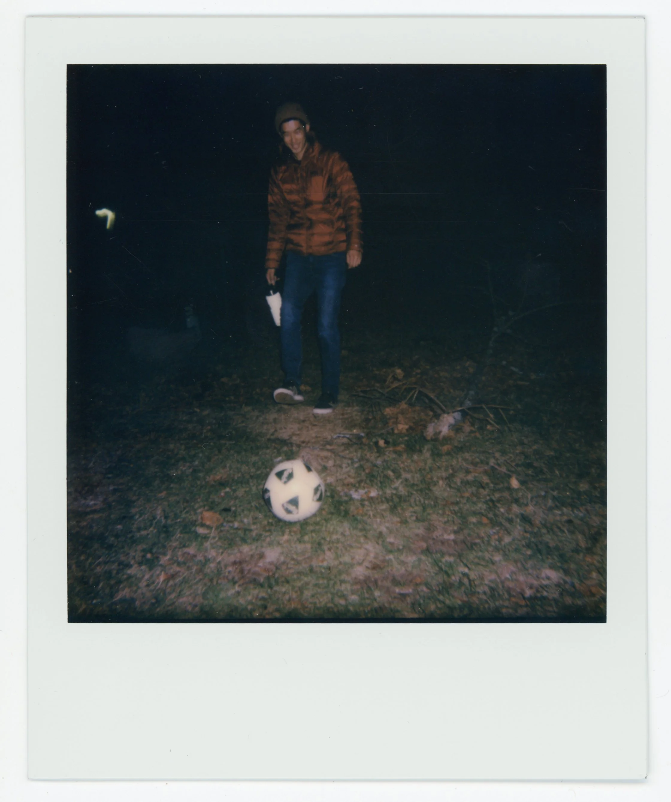 Person standing outdoors at night with a soccer ball on the ground in front of them.