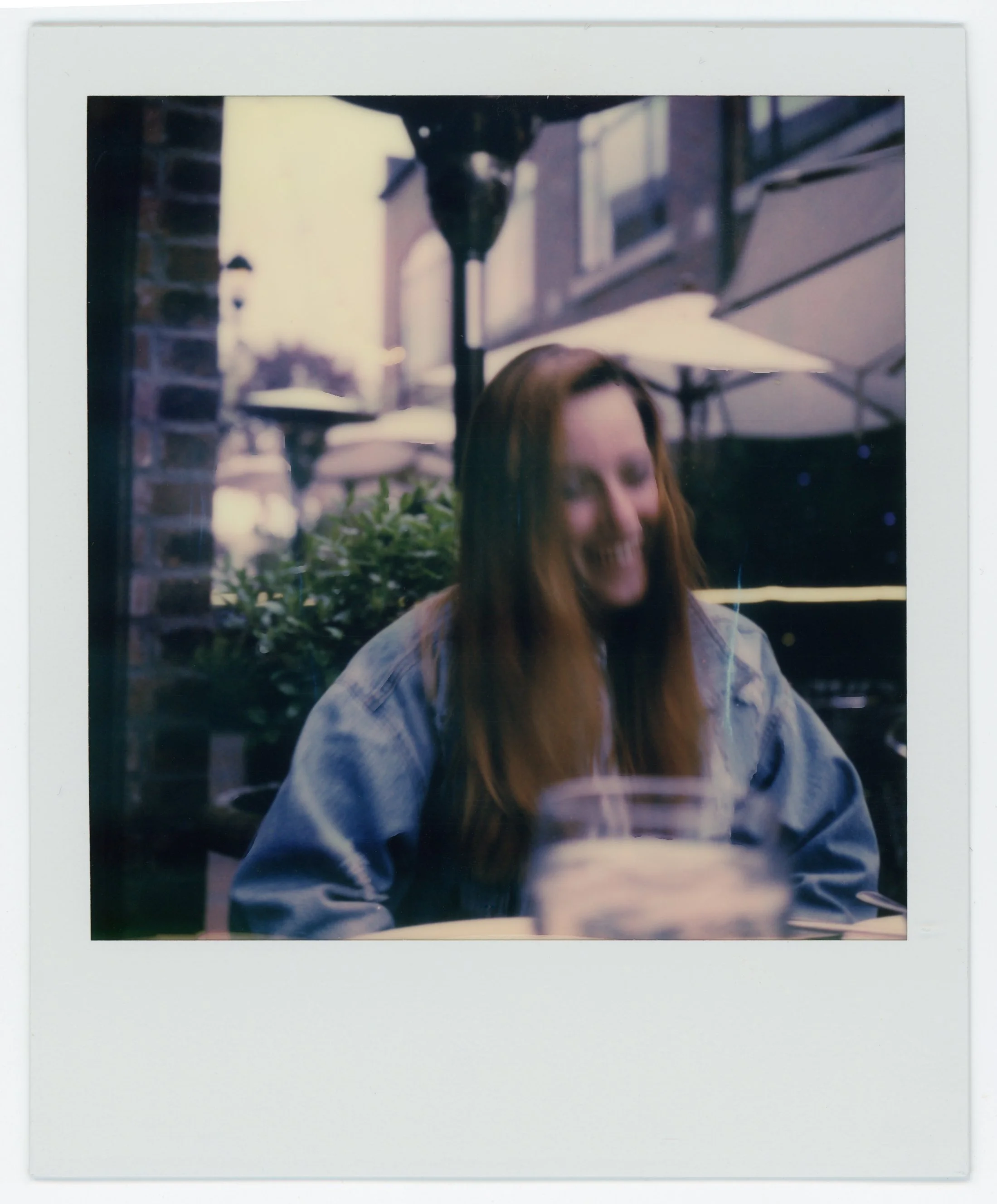 A woman with long red hair smiling and sitting at an outdoor table in a café or restaurant, with umbrellas and buildings in the background.