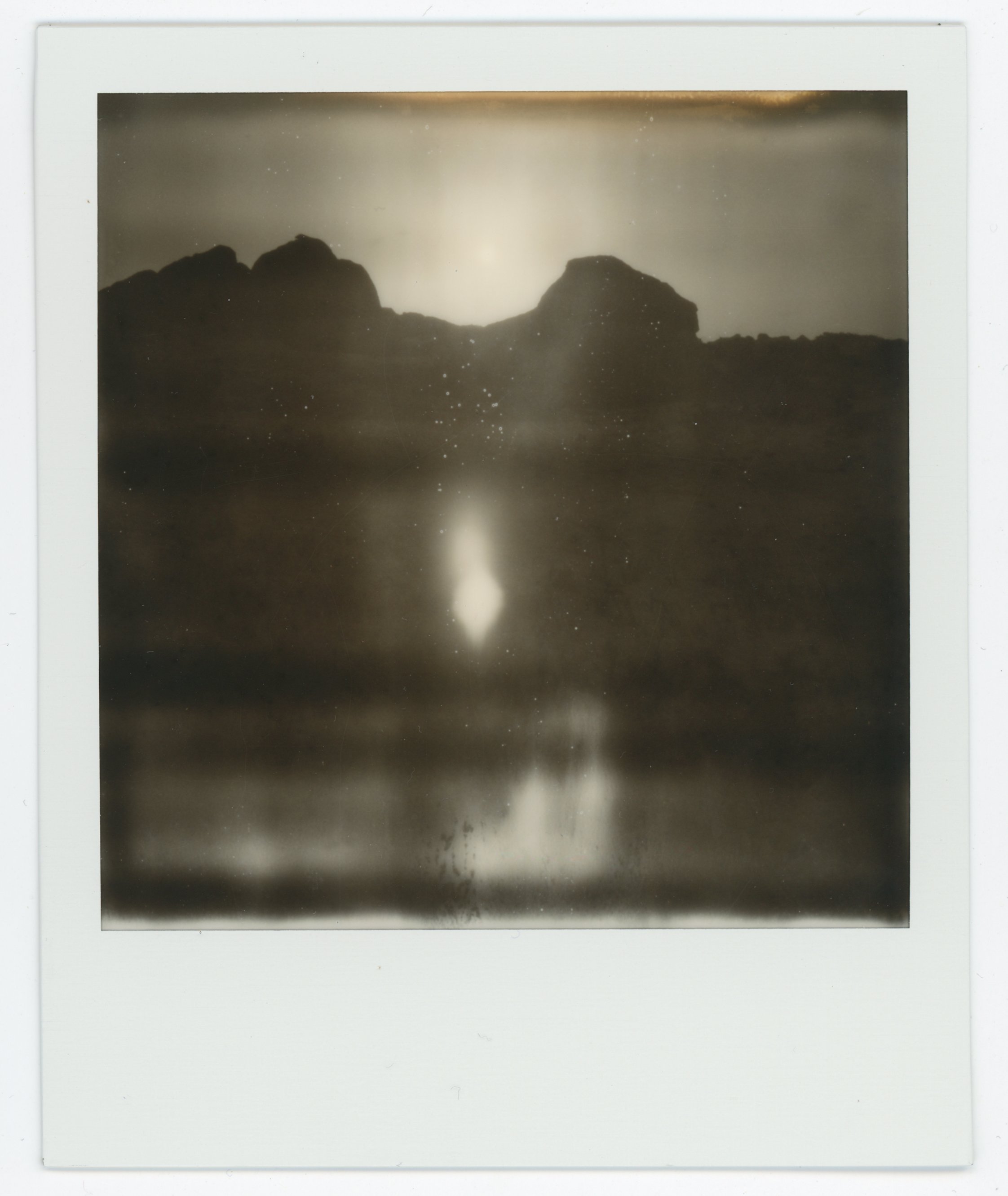 Black and white Polaroid photo of a mountainous landscape reflected on a body of water, with a bright glow and star-like points in the sky.