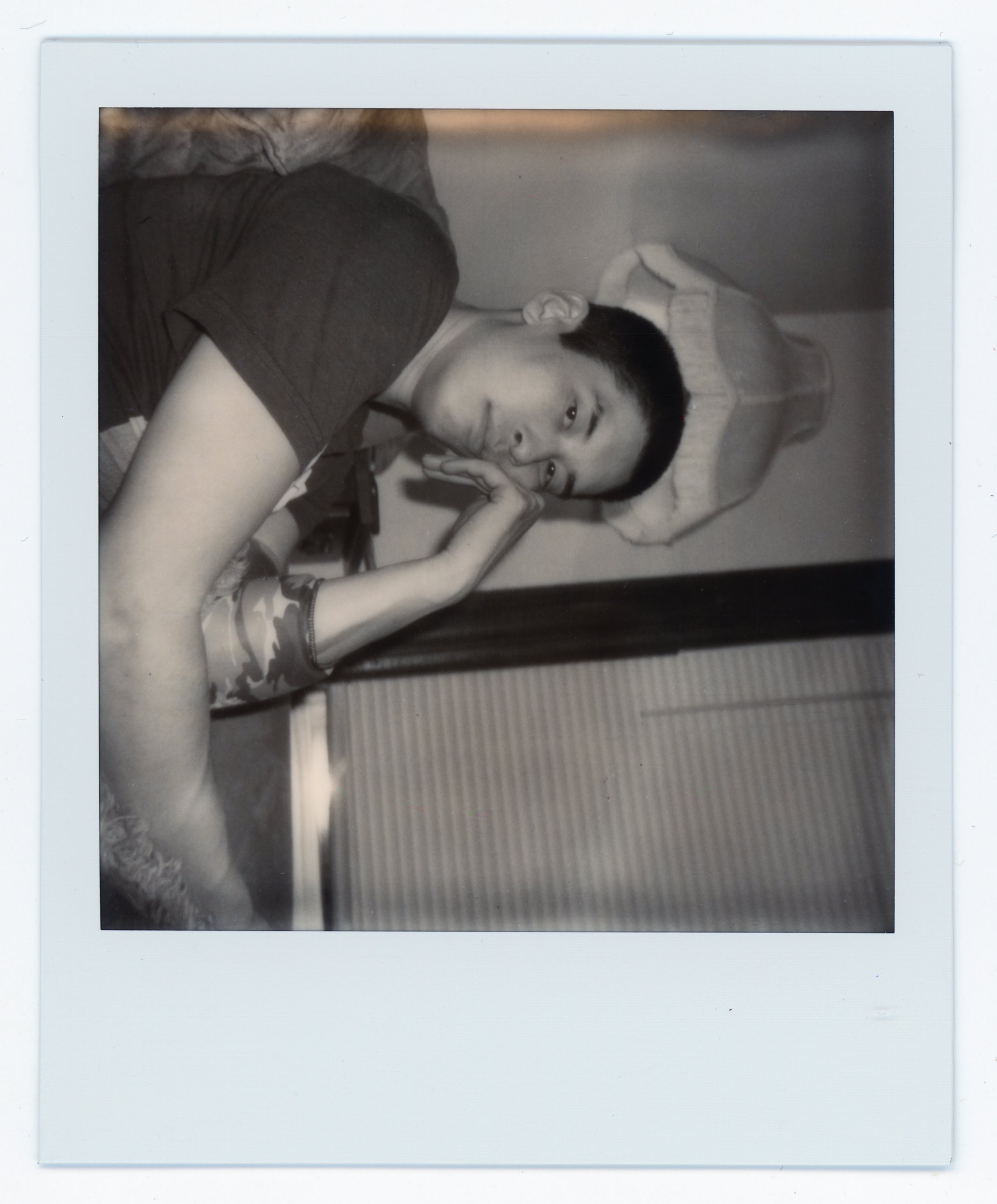 A young person with short hair lying on a bed, resting their head on their hand, looking towards the camera. The photo is in black and white.