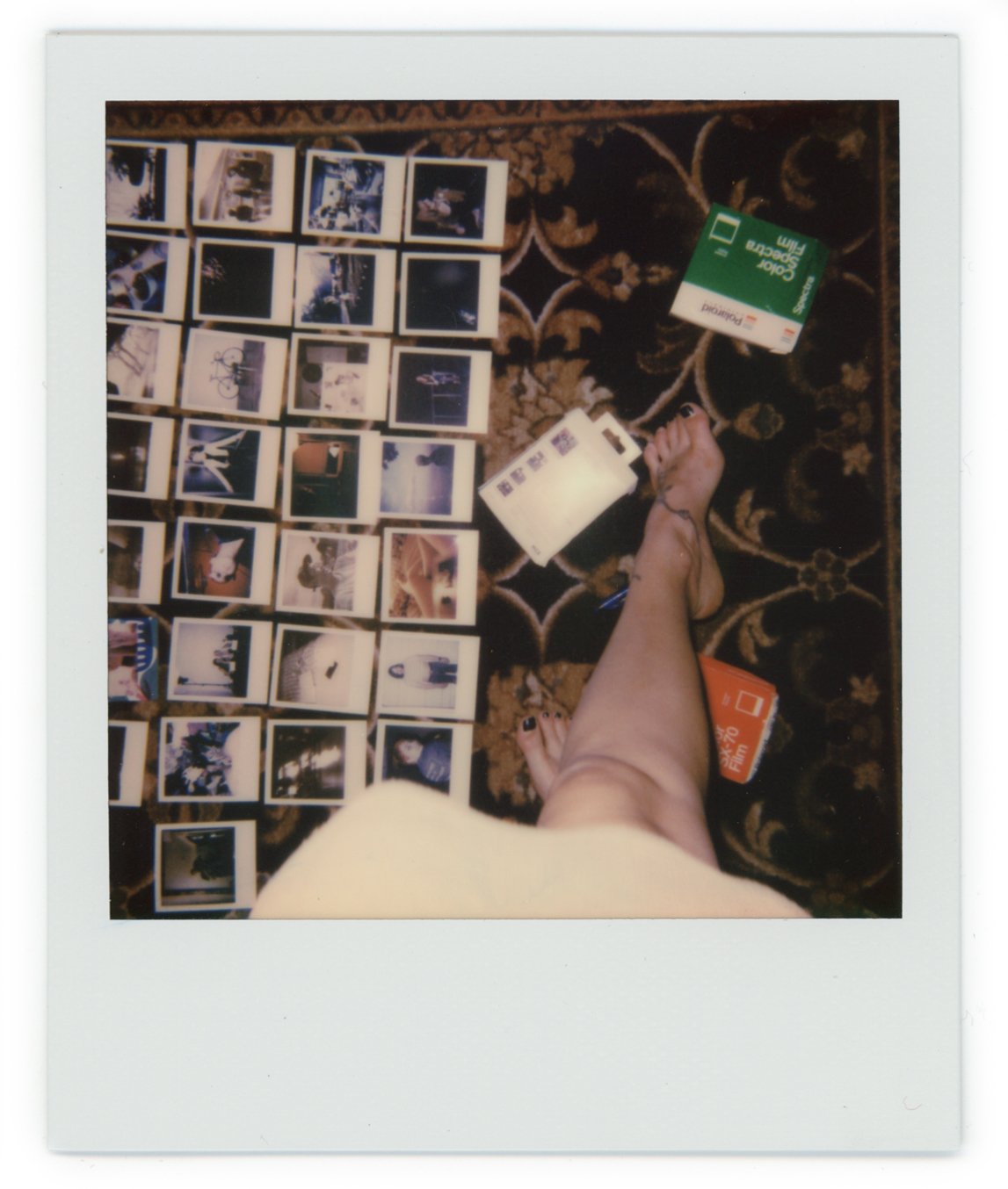 Polaroid photographs laid out on a patterned carpet with a person's lying foot in the foreground, with a green card and a white peripheral device.