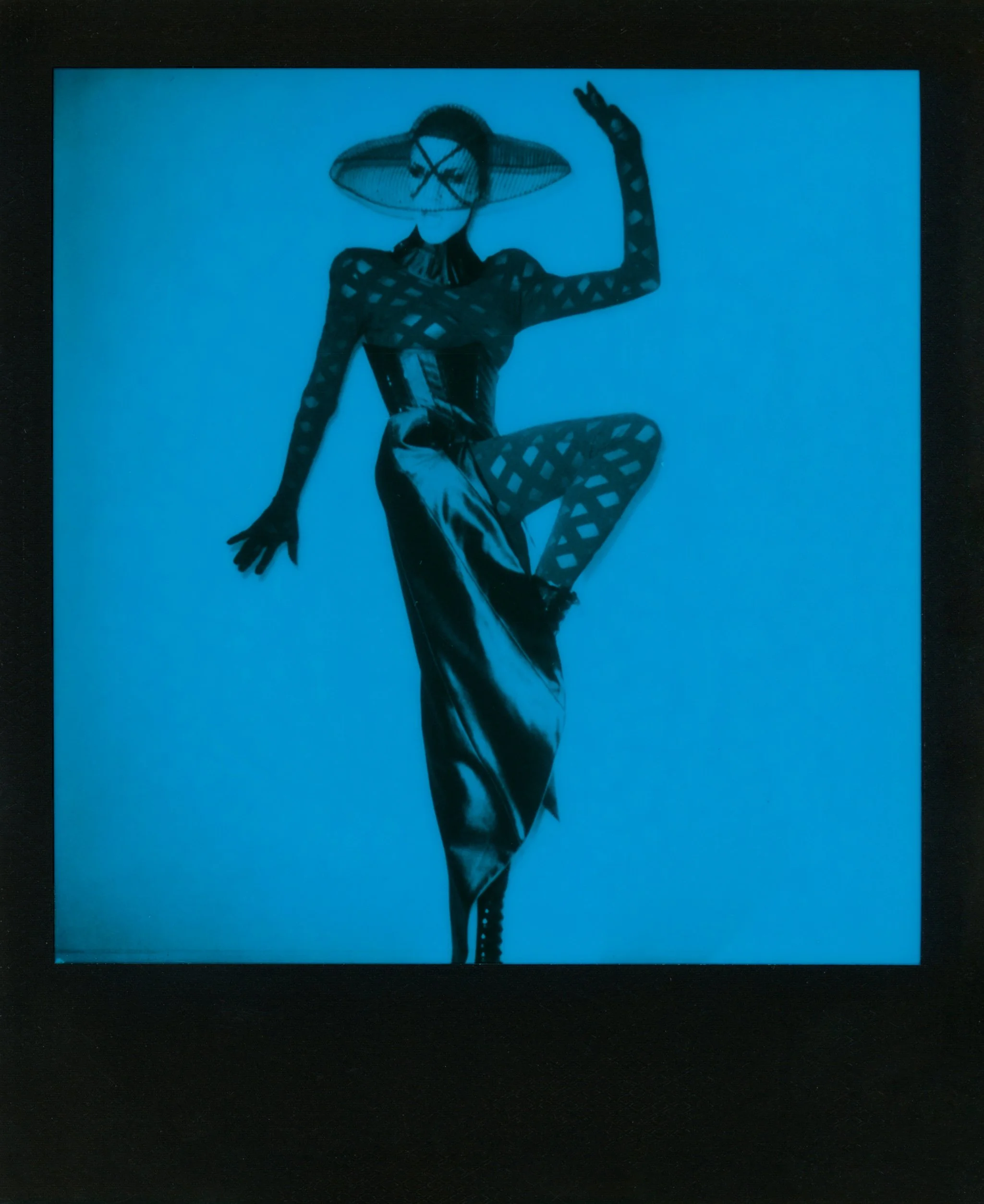 A woman in black fishnet stockings, a corset, and a wide-brimmed hat posing against a plain background.
