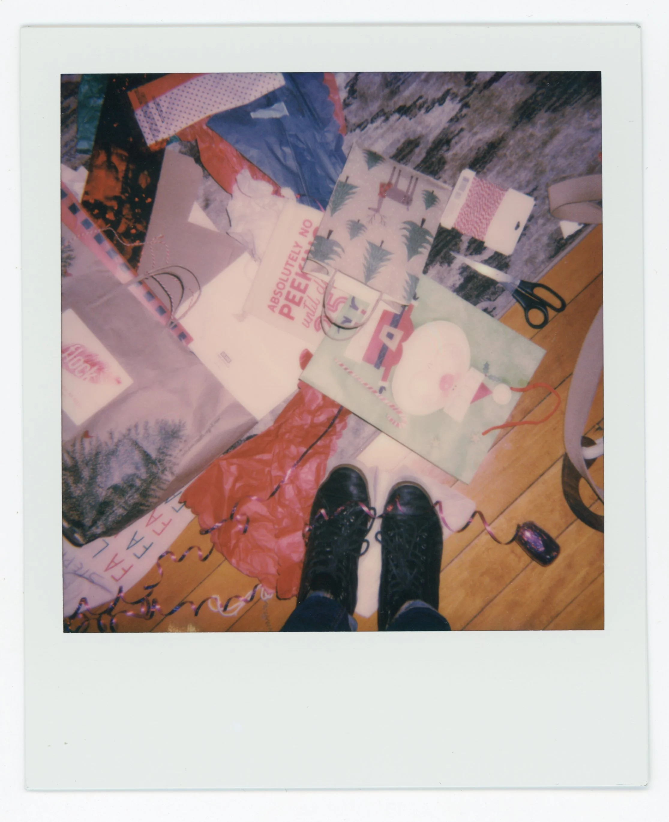 Photo taken from above showing a person wearing black boots standing on a wooden floor, surrounded by wrapping paper, scissors, and decorations, indicating gift wrapping activity.