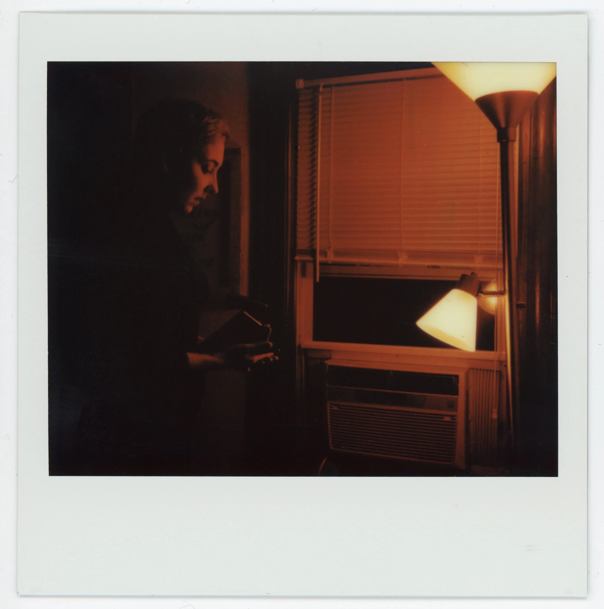 A woman stands in a dimly lit room at night, looking at her phone with a window and a lit floor lamp in the background.