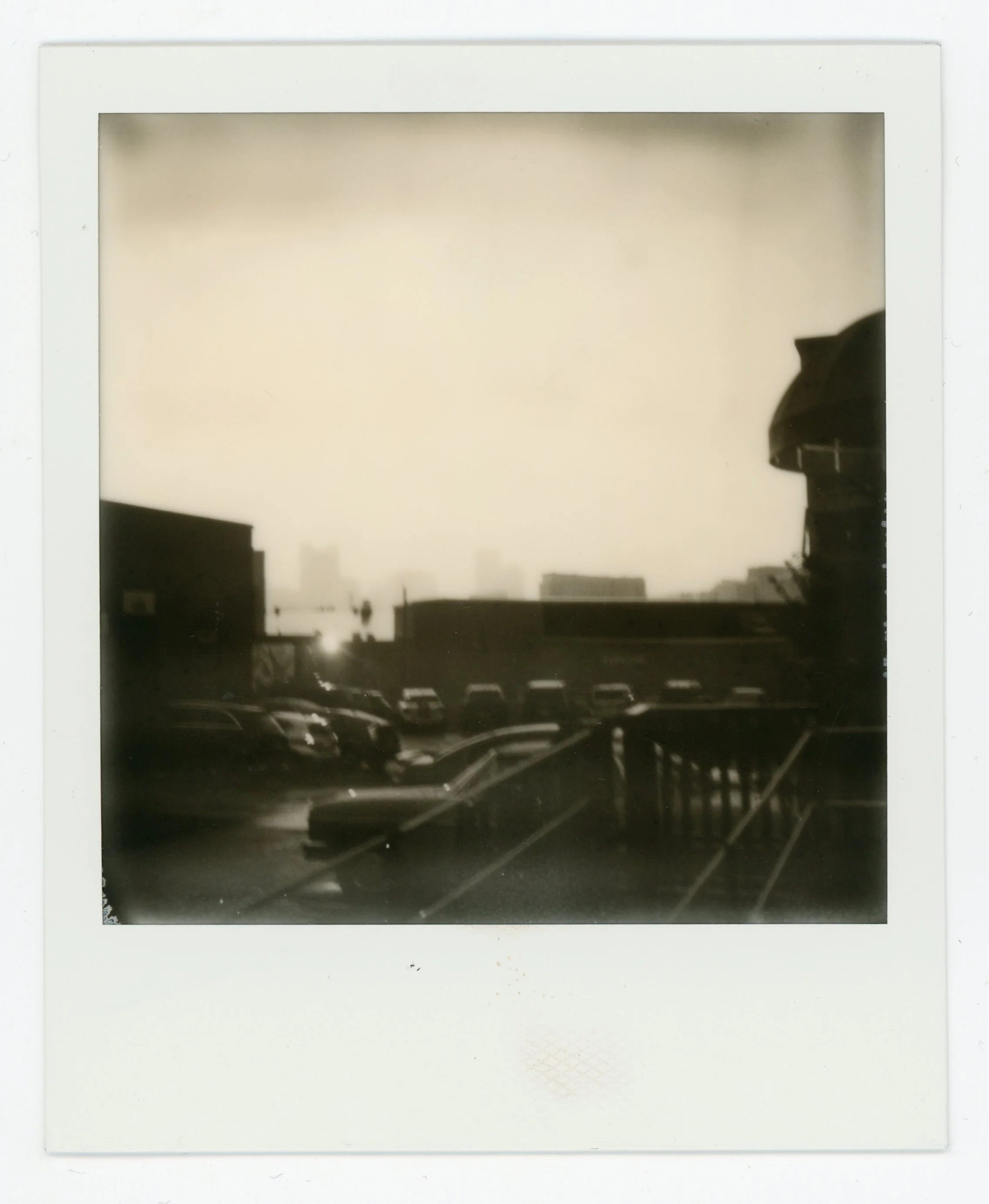 Black and white Polaroid photo of a cityscape with parked cars in the foreground, buildings, and a setting sun in the background.