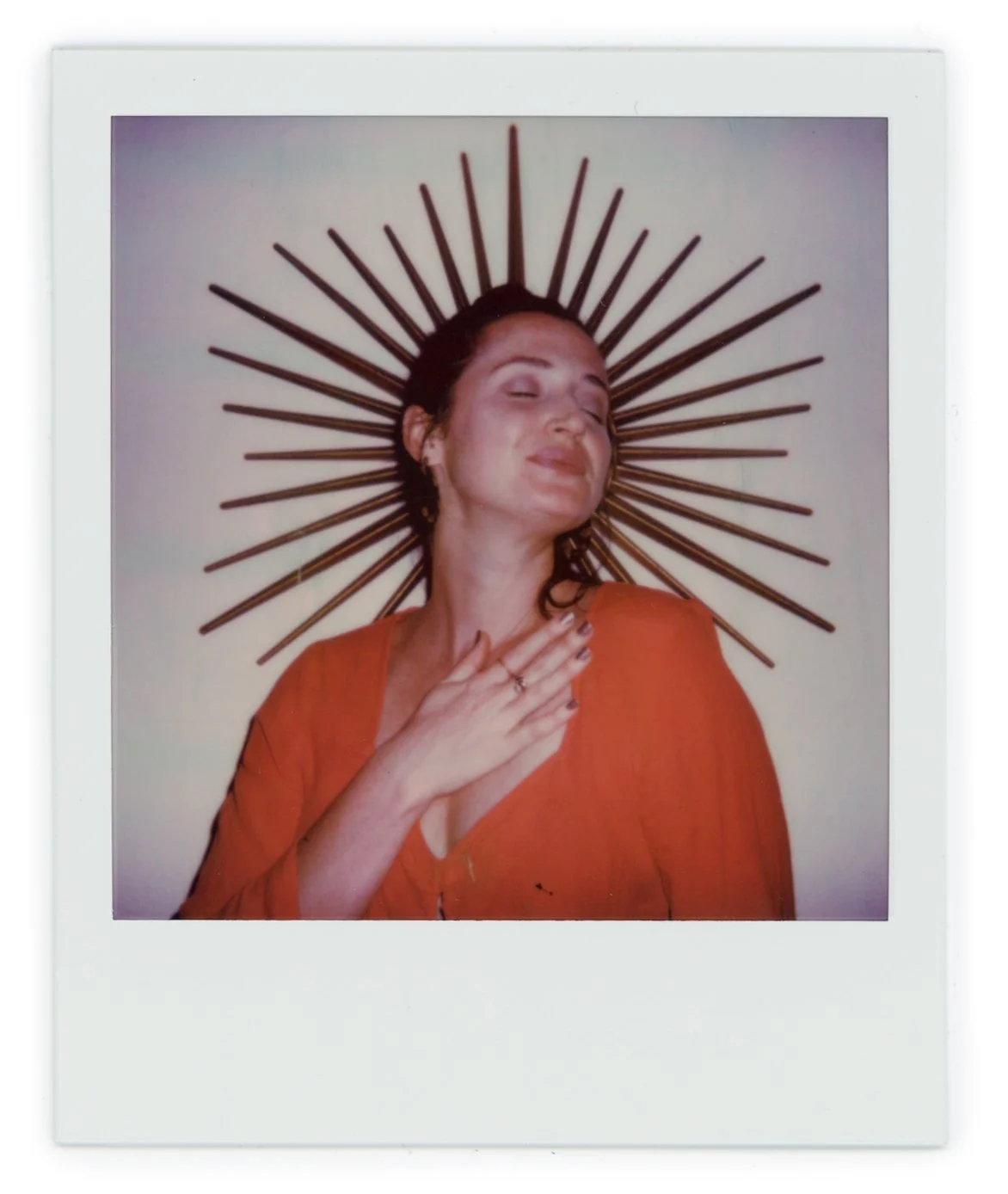 Woman with dark hair in a burnt orange top, wearing a halo-like sunburst crown behind her head, smiling with eyes closed and hand on her chest.