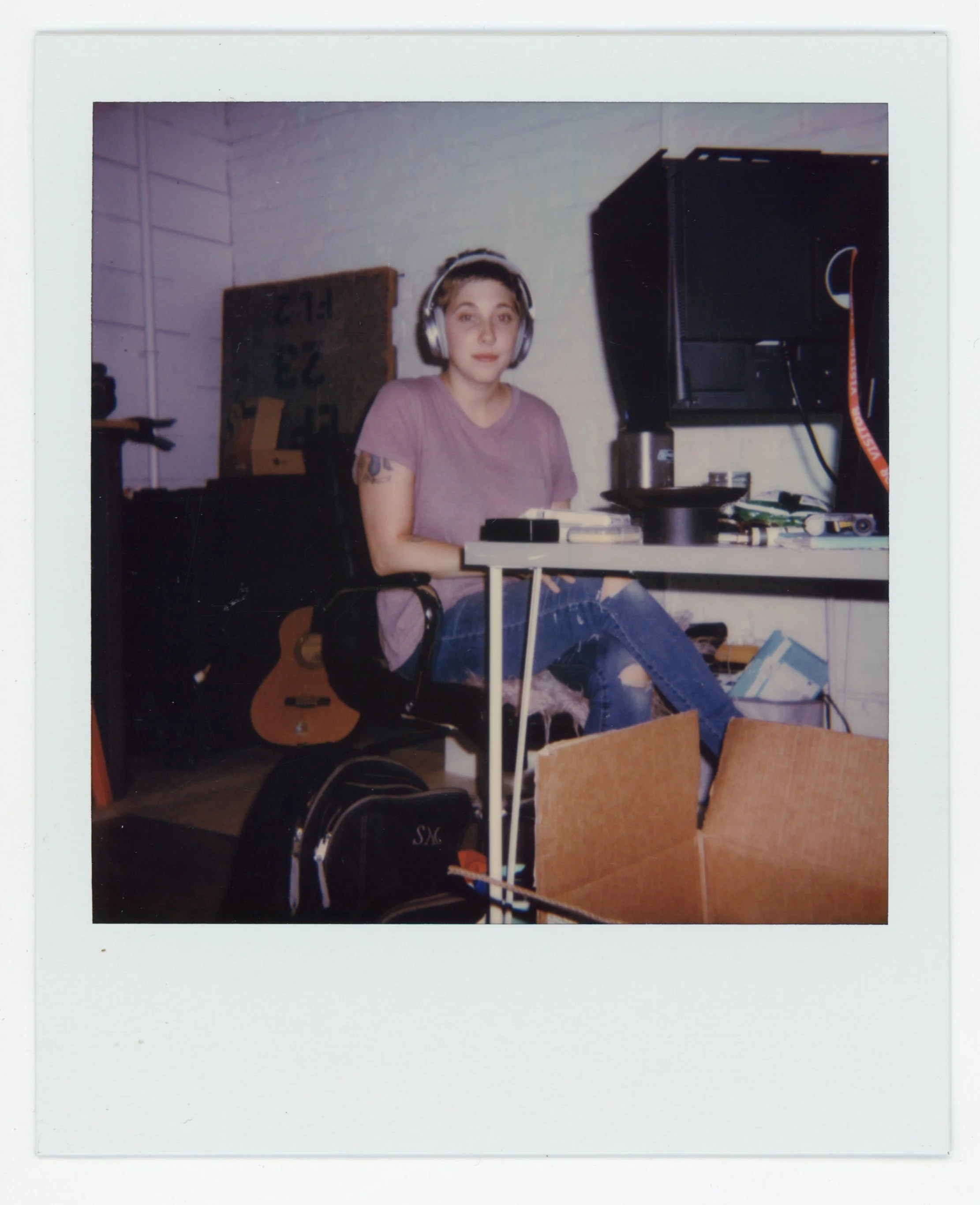 Young woman sitting at a desk with headphones, a guitar behind her, and a cardboard box in front, in a room with a white brick wall.