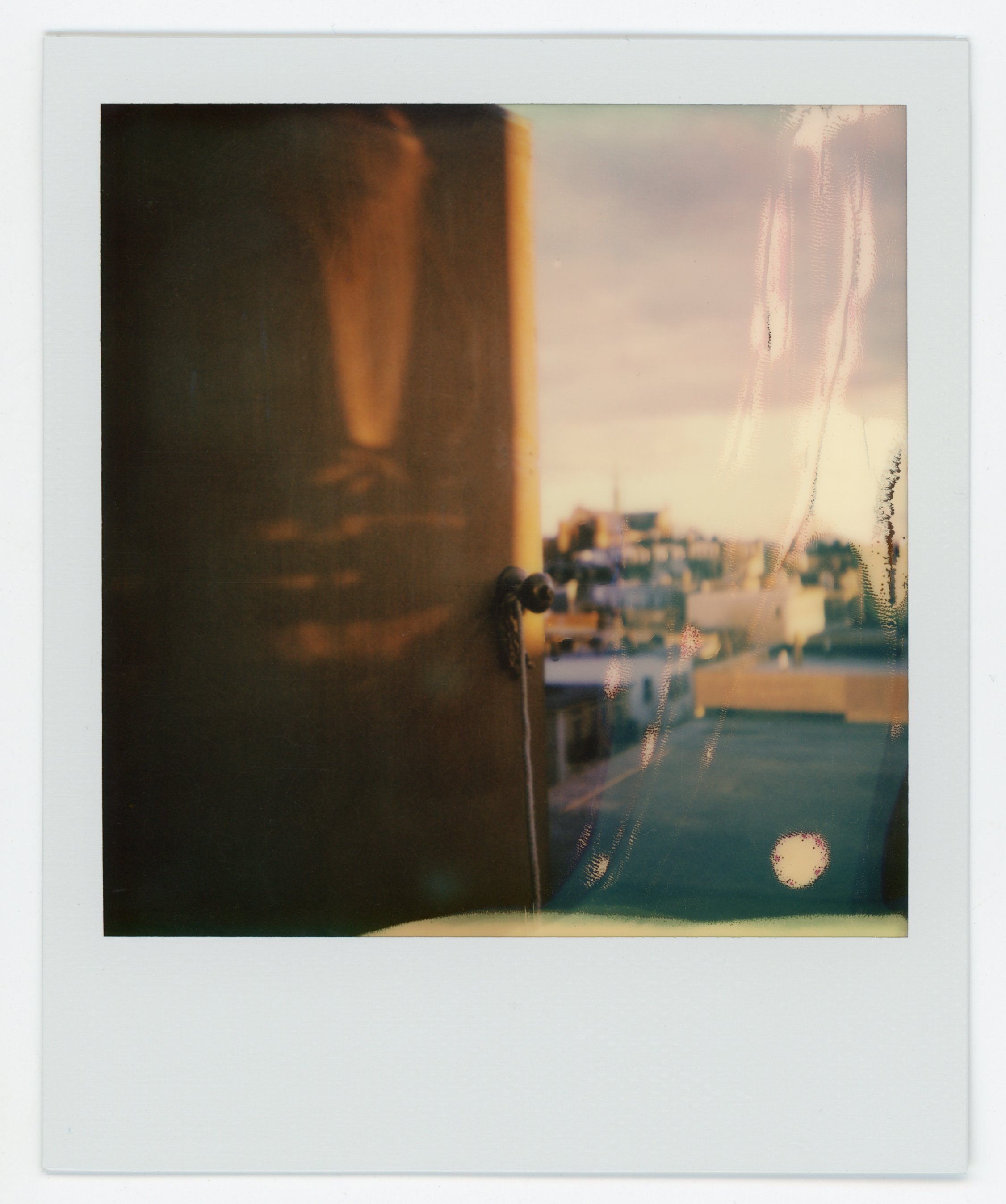 Polaroid photo of a window with a cityscape view in the background and a wooden door partially blocking the view.