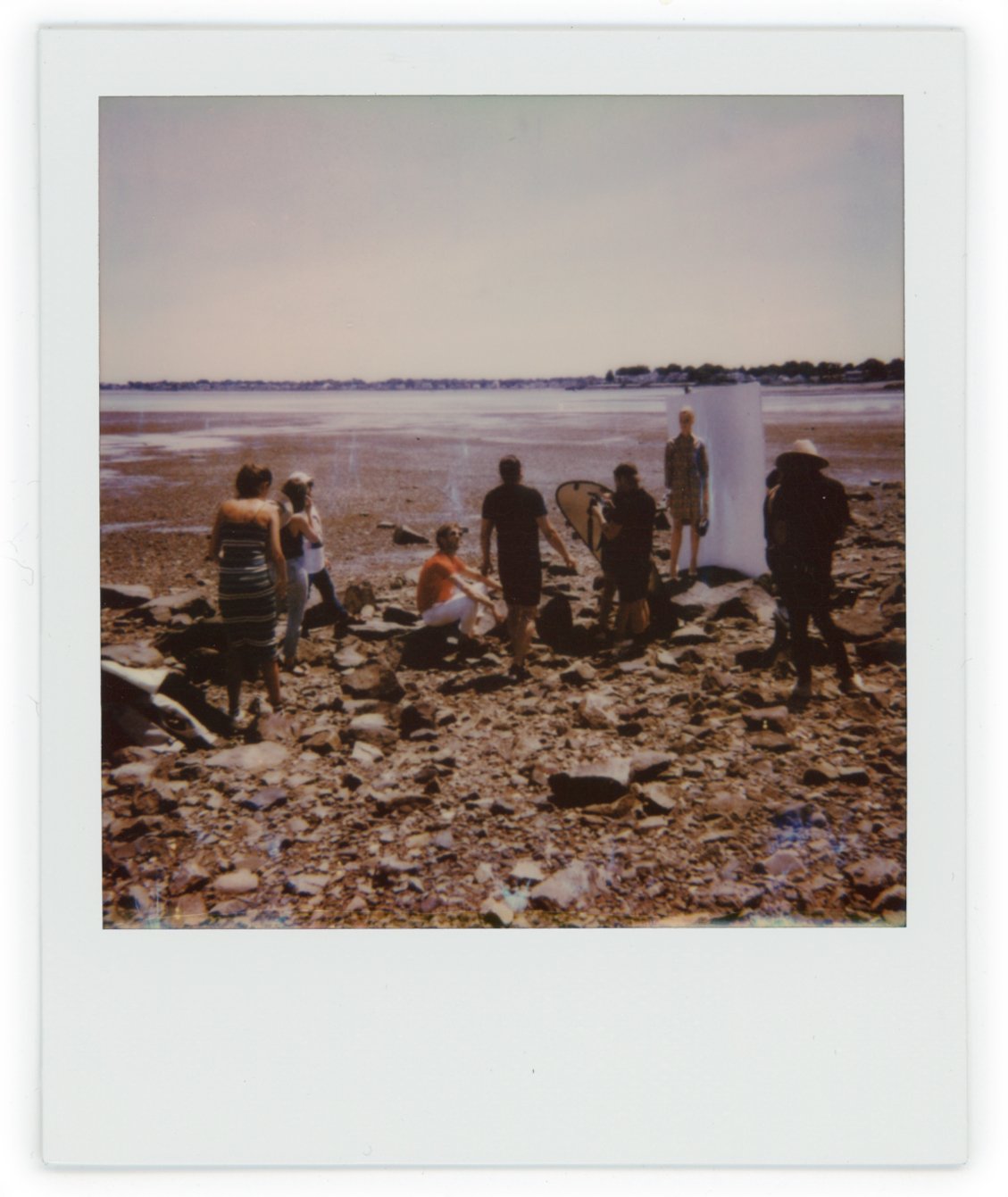 Group of people on a rocky beach near the water, with some standing around a white backdrop and photography equipment, under a clear sky.