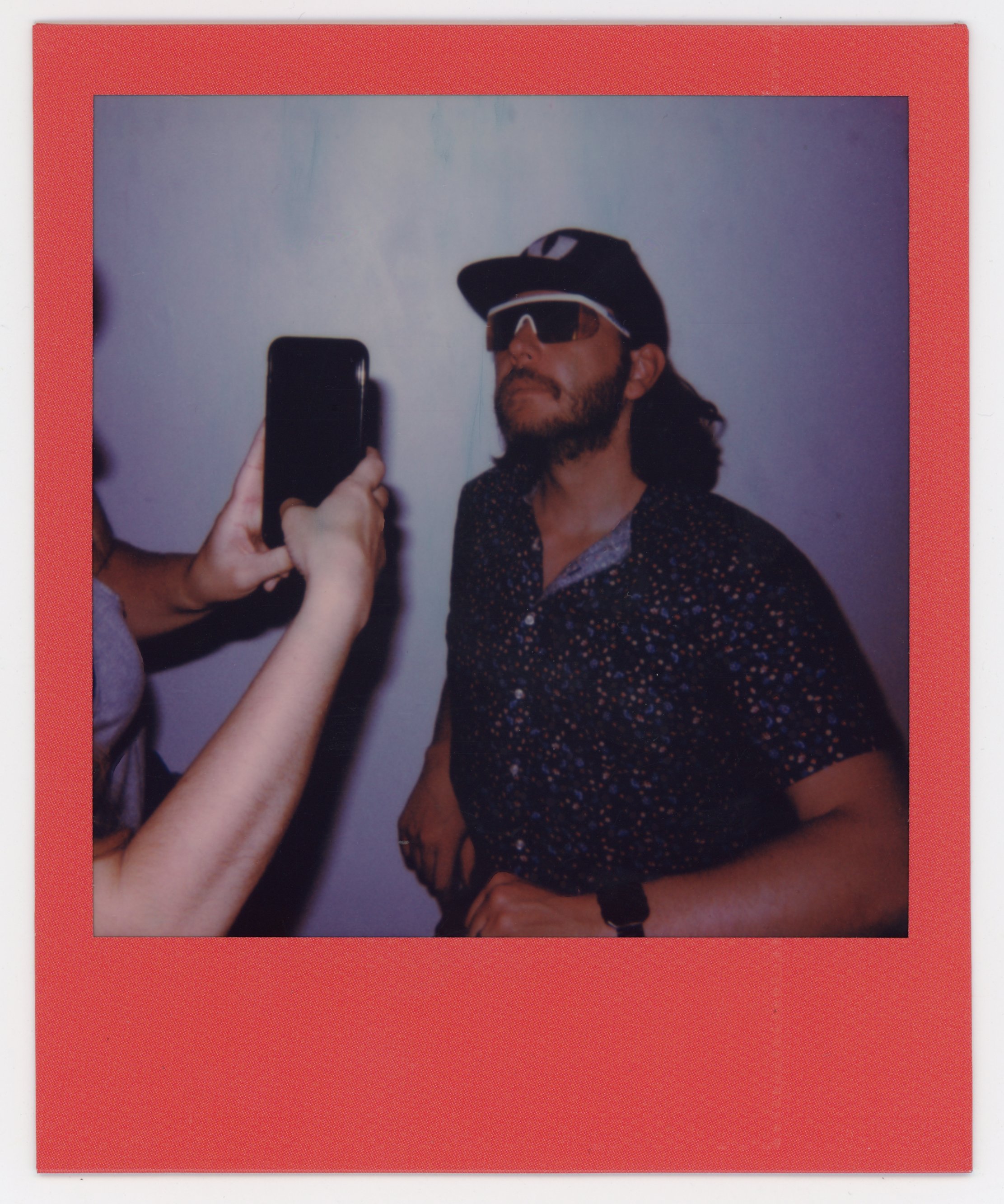A man posing for a photo, wearing sunglasses, a cap, and a patterned shirt, while someone else takes a picture with a smartphone.