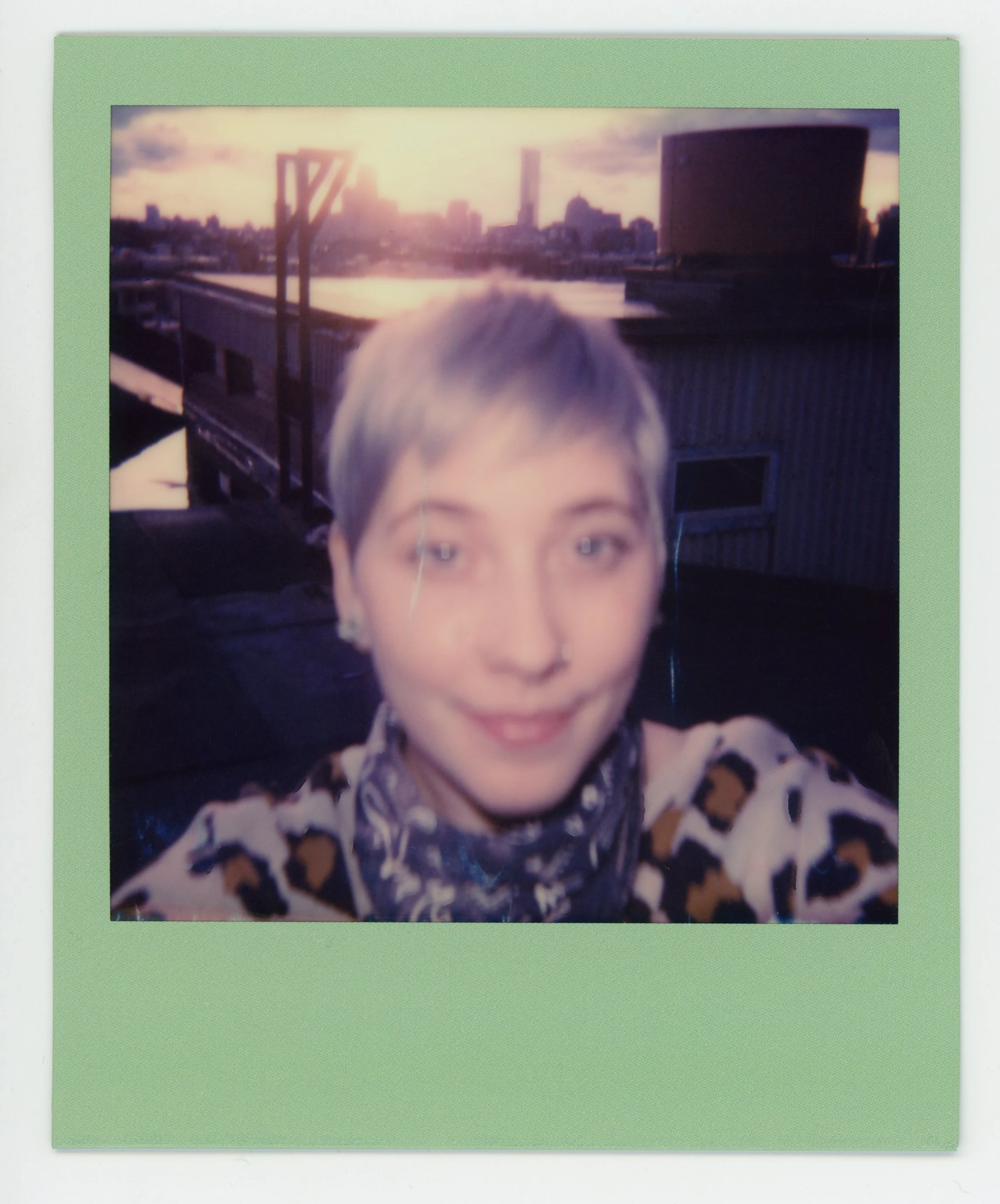 A woman with short blonde hair taking a selfie on a rooftop with a city skyline and water in the background during sunset or sunrise.