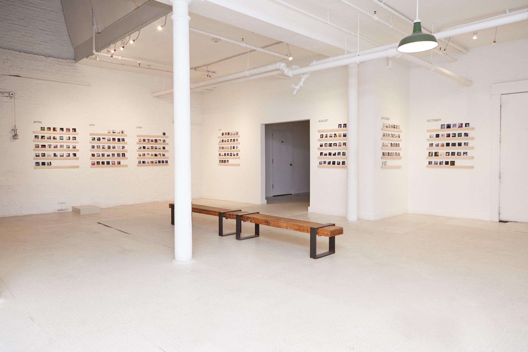 Art gallery with photo displays arranged in monthly sections on white walls, a wooden bench in the foreground, white walls, exposed pipes, and modern lighting.